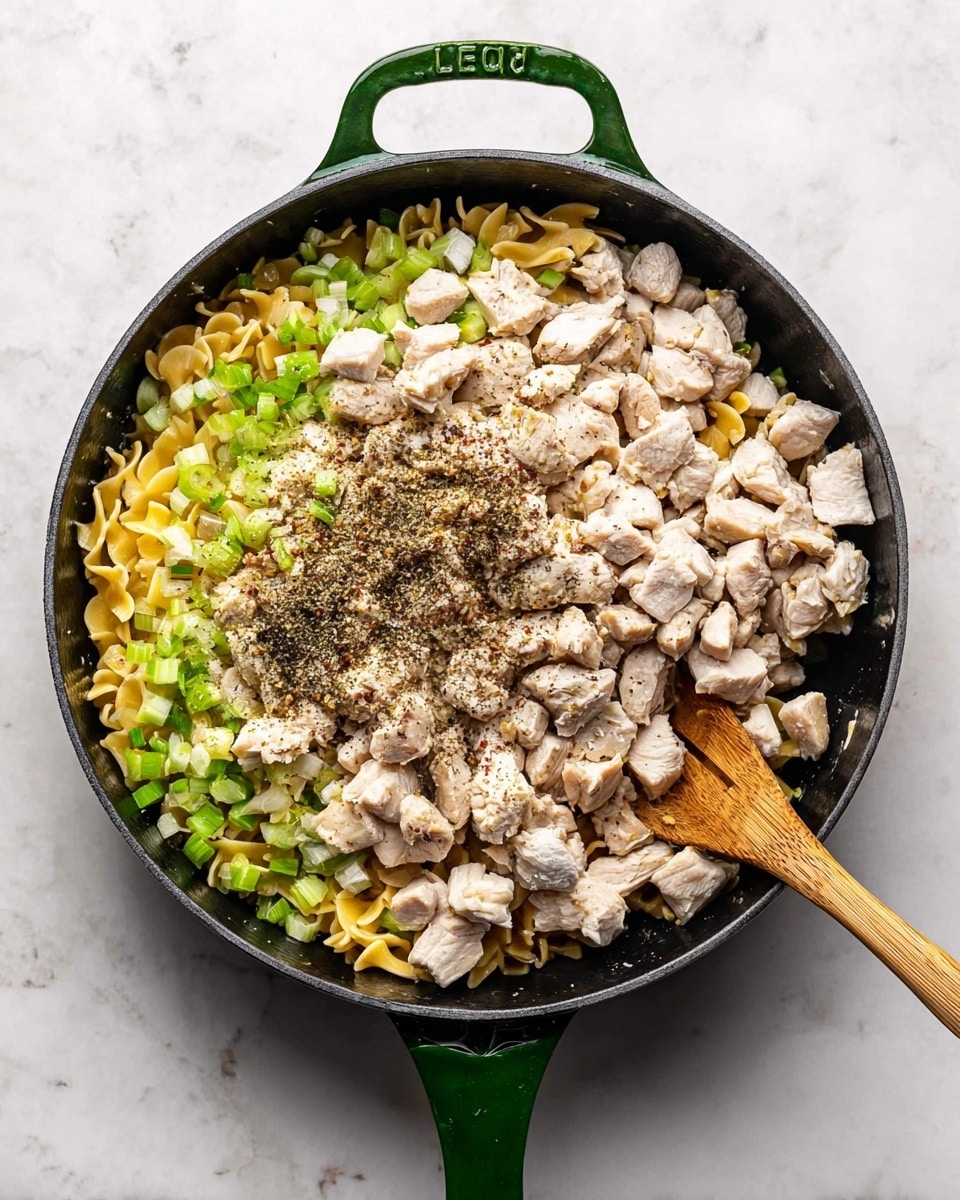 A black skillet with a green handle is placed on a white marbled surface, filled with layered ingredients. The bottom layer consists of yellow pasta pieces mixed with chopped green celery, creating a textured base. On top, there is a thick layer of small, cubed light beige chicken pieces scattered unevenly. Dark brown dried herbs and black pepper are sprinkled generously over the chicken. A wooden spoon rests inside the skillet, partly under the chicken, ready to mix the ingredients. Photo taken with an iphone --ar 4:5 --v 7