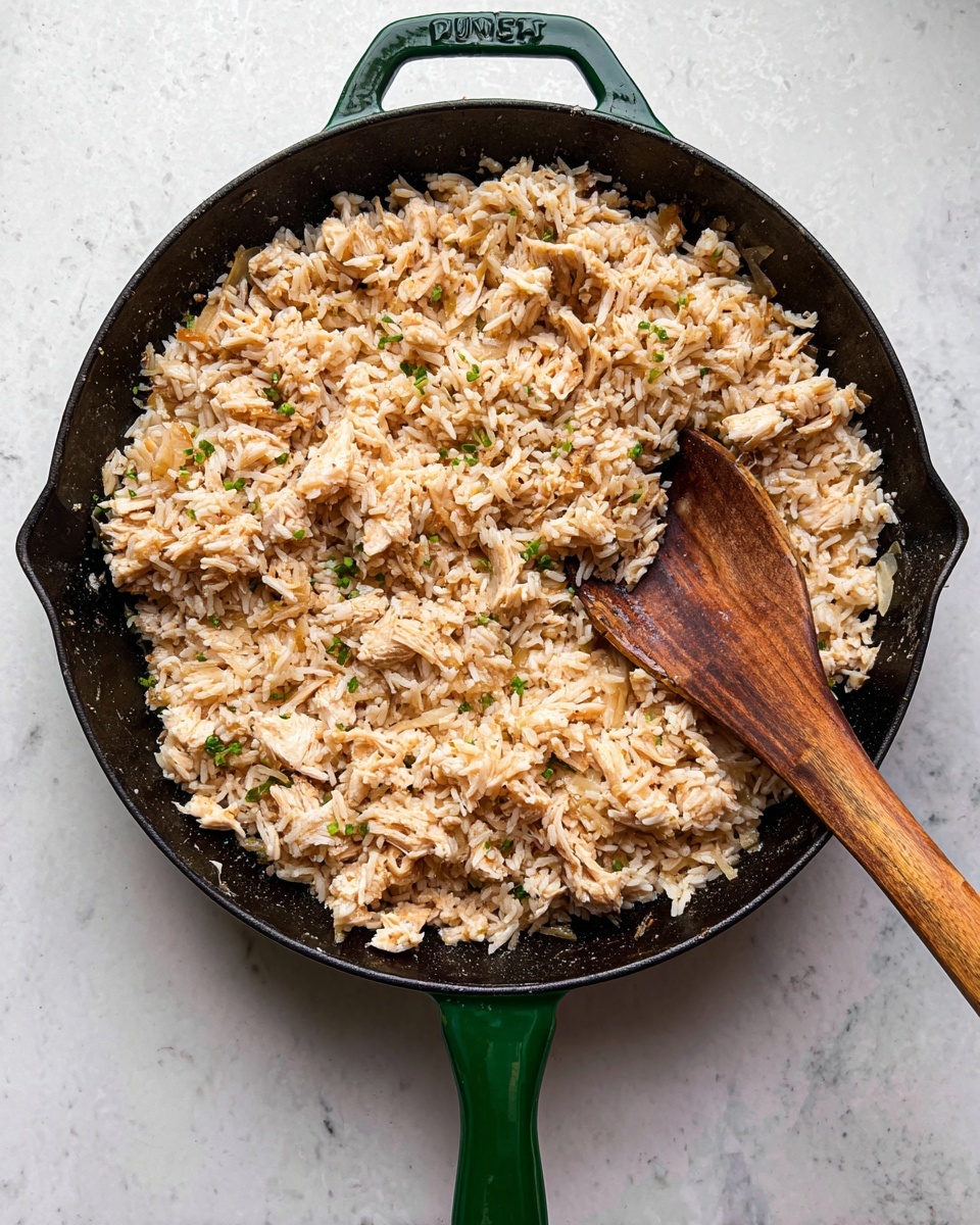 A black cast iron skillet with a green handle holds a single layer of cooked rice mixed with small chunks of light brown cooked chicken pieces and light-colored onions. The rice and chicken mixture looks moist with a slightly uneven texture. A wooden spoon with a long handle lays inside the skillet, partly resting on the food. The skillet sits on a white marbled surface. photo taken with an iphone --ar 4:5 --v 7