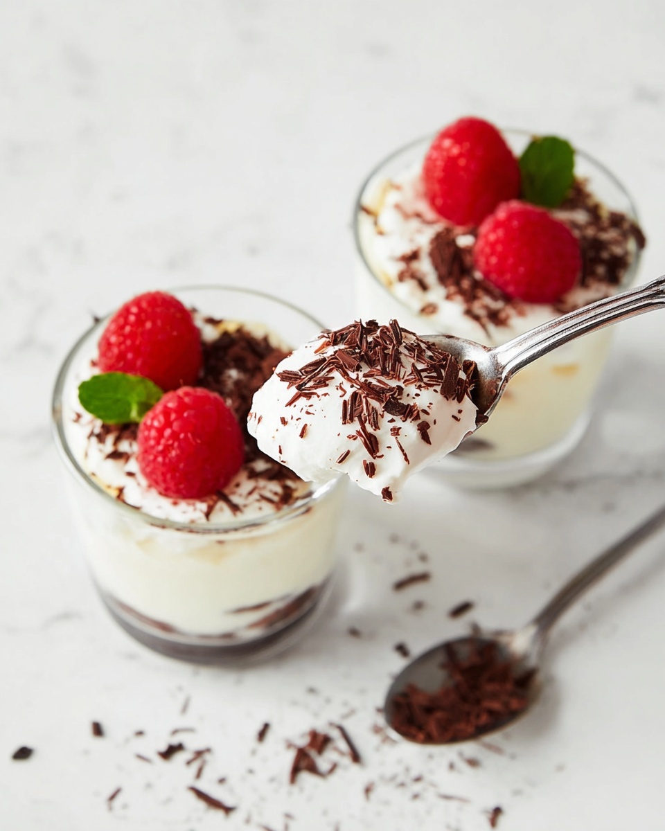 The image shows two glass cups filled with a three-layer dessert. The bottom layer is light cream. The middle layer is white cream topped with dark chocolate shavings sprinkled unevenly. On top of each dessert, two red raspberries sit close together, with a small green leaf for decoration. In the front, a spoon with white cream and chocolate shavings is held above one cup, showing texture clearly. The background is a white marbled texture with some scattered chocolate shavings and a spoon resting beside the cups. Photo taken with an iphone --ar 4:5 --v 7