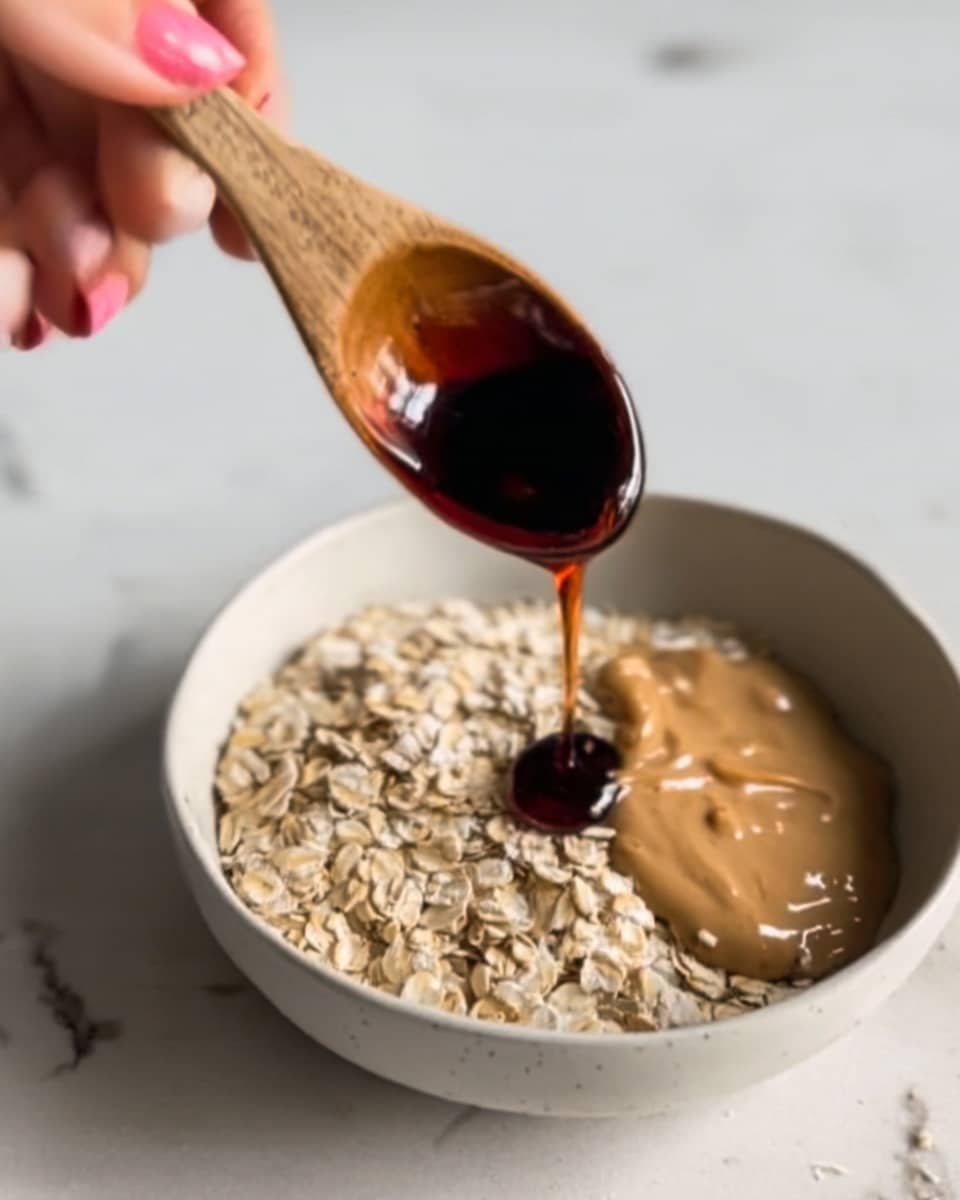 A close-up image shows a wooden spoon held by a woman's hand pouring dark brown liquid, likely maple syrup, over a white bowl filled with light beige rolled oats and a smooth, light brown peanut butter layer on one side. The bowl sits on a white marbled surface, and the focus is on the pouring action with soft, natural light highlighting the textures of the oats and peanut butter. Photo taken with an iphone --ar 4:5 --v 7