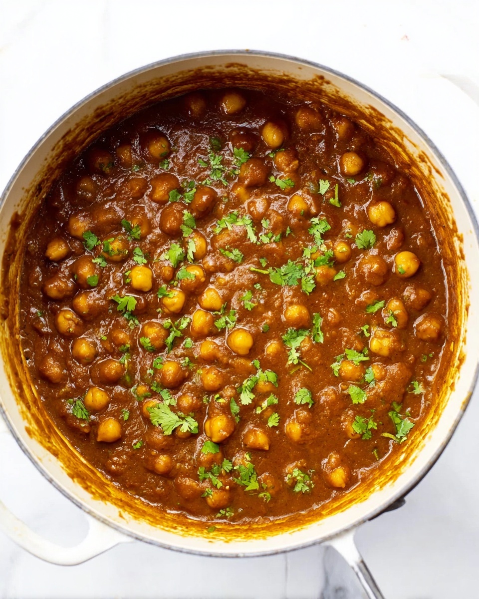 The image shows a white pot filled with rich brown chickpea curry, thick in texture, with whole chickpeas visible throughout the sauce. Bright green chopped cilantro leaves are sprinkled on top, adding a fresh contrast to the warm brown color. The pot sits on a white marbled surface, and some sauce stains can be seen on the inside edges of the pot. The overall look is hearty and inviting, with the colors and textures making the dish look freshly cooked and ready to serve. photo taken with an iphone --ar 4:5 --v 7