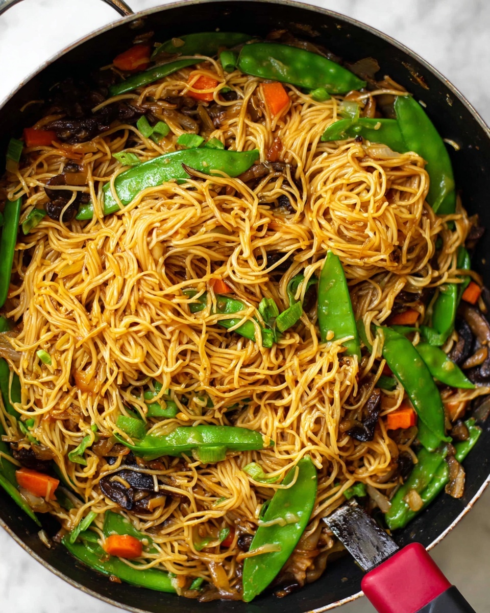 This image shows a pan filled with stir-fried noodles that are light brown and soft, mixed with bright green snap peas scattered throughout. Along with the noodles, there are small orange carrot pieces and slices of dark brown mushrooms. The noodles form the top layer, loosely curled and piled unevenly. The snap peas and carrot pieces are spread around and under the noodles, adding pops of color and a fresh texture. The pan is black with a handle that has a red and black grip, placed on a white marbled surface. Photo taken with an iphone --ar 4:5 --v 7