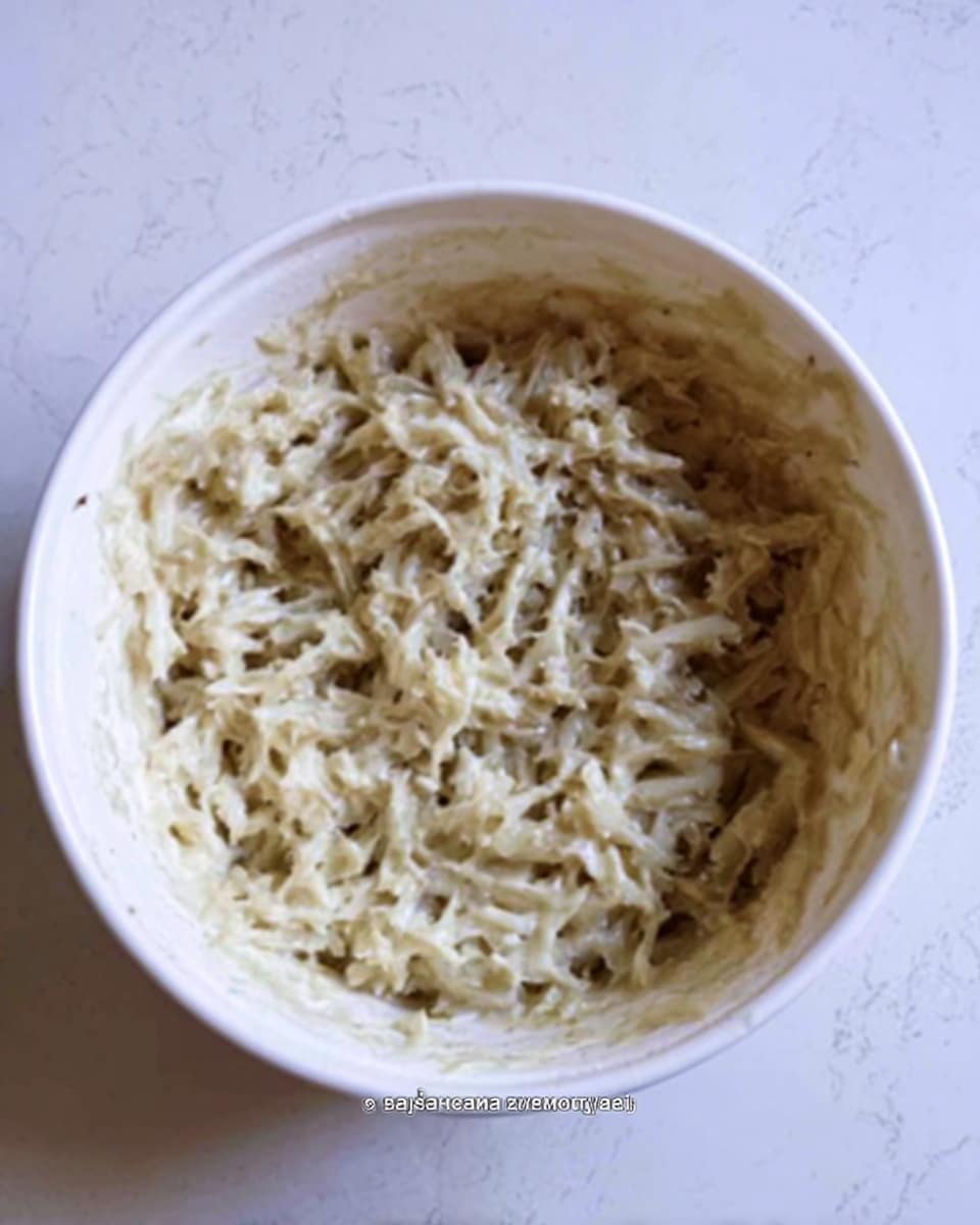 A white bowl filled with a creamy mixture that looks like small shredded pieces combined with a pale sauce, with a few darker spots throughout the mixture. The texture appears wet and soft, with the contents evenly spread inside the bowl. The bowl is placed on a white marbled surface. Photo taken with an iphone --ar 4:5 --v 7