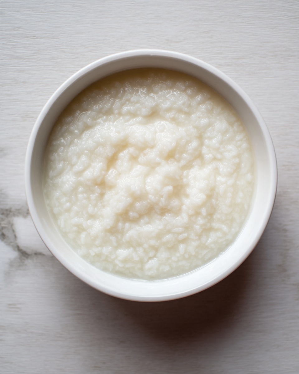 A white bowl filled with soft, thick white rice porridge that looks creamy and smooth with small, tender rice grains visible throughout the surface; the bowl is placed on a white marbled surface, creating a clean and simple background. Photo taken with an iphone --ar 4:5 --v 7