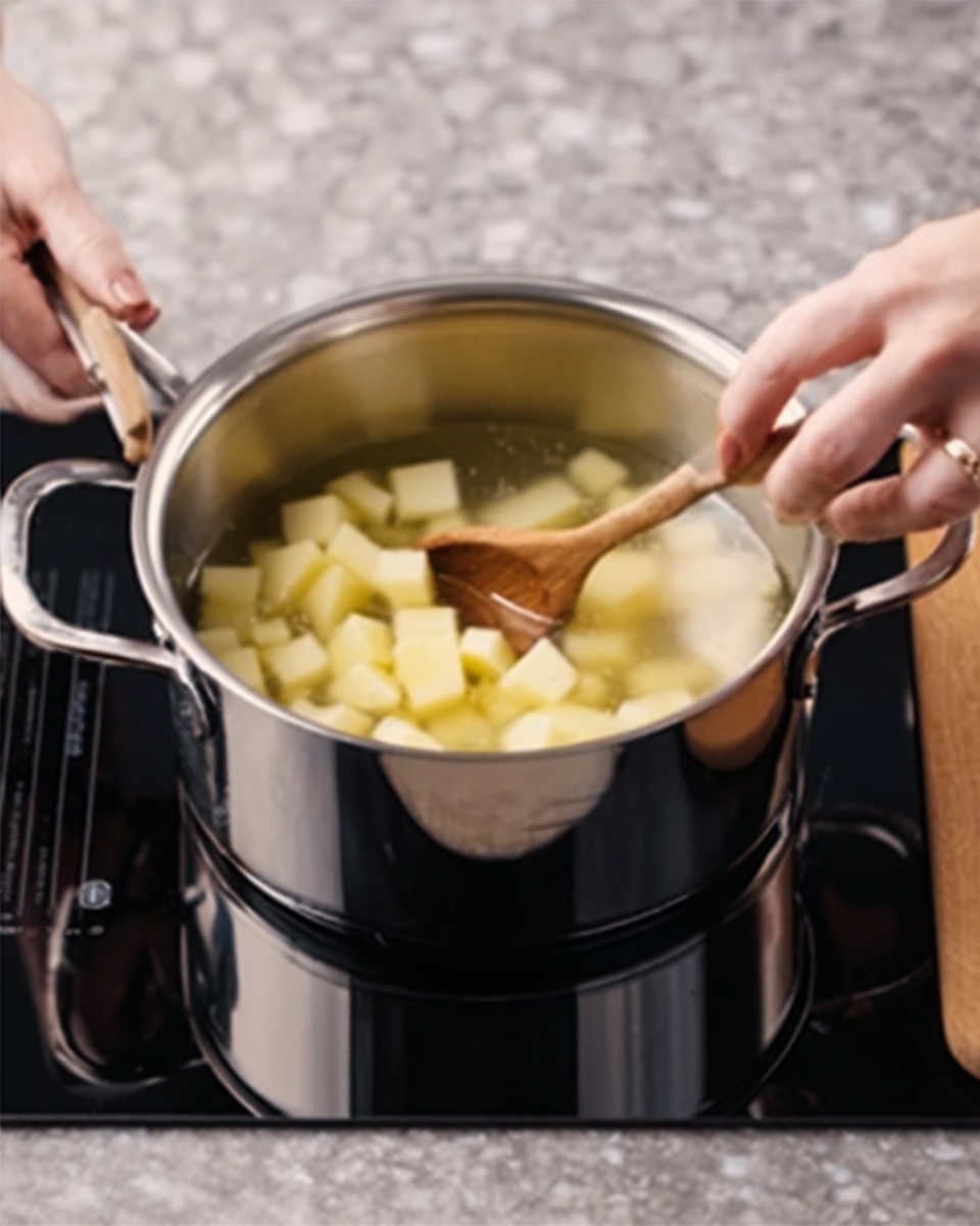 A stainless steel pot sits on a black stove top with a white marbled background. Inside the pot, light yellow potato cubes fill the first layer, topped by a layer of water covering them. A woman's hand holds a wooden spoon stirring the potatoes gently from top right, while another woman's hand holds the pot's handle on the left side. The scene focuses on the cooking process with clear reflections on the shiny pot surface. Photo taken with an iphone --ar 4:5 --v 7