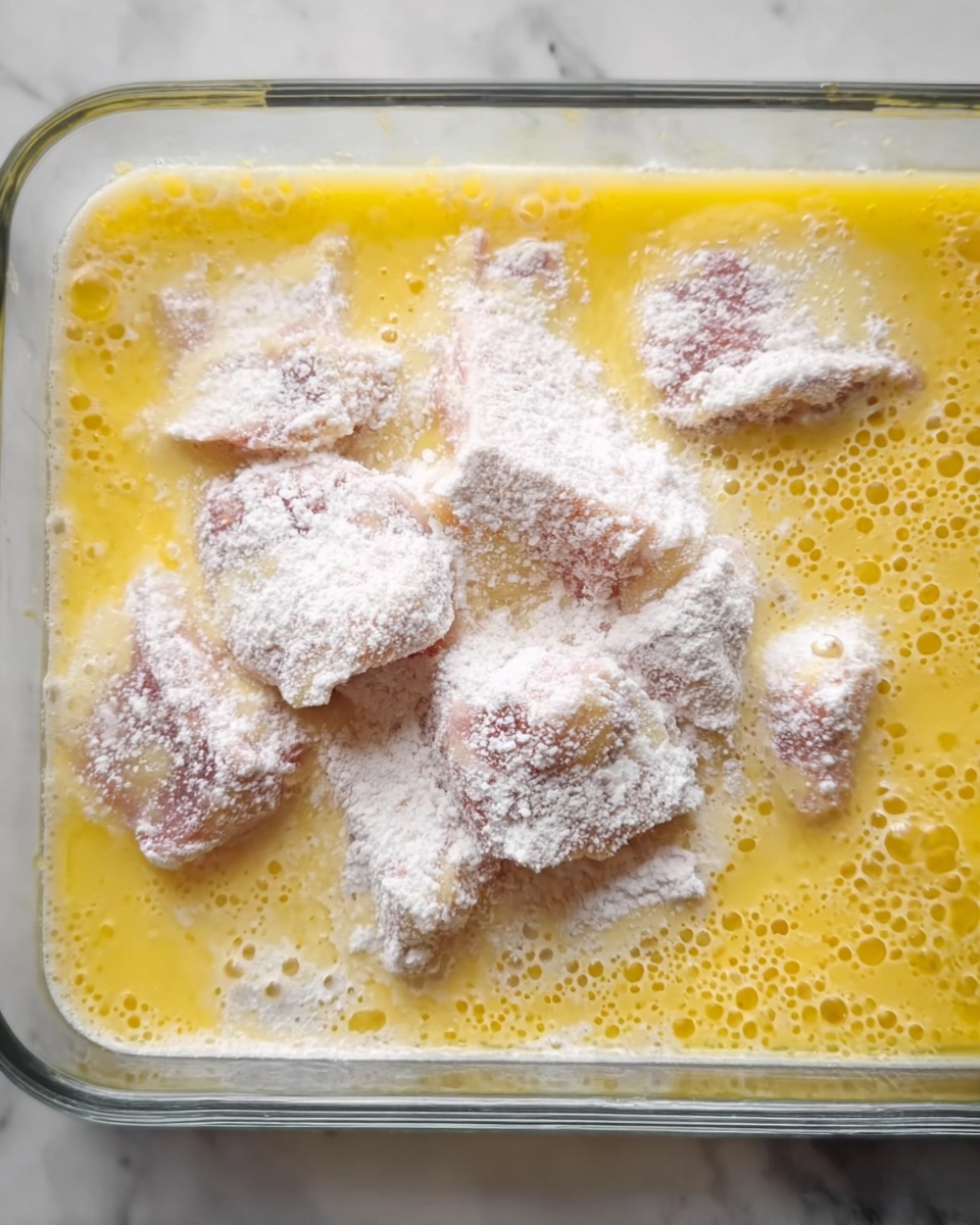 The image shows raw pieces of meat covered with white flour, placed inside a clear glass container with a yellow liquid mixture, likely beaten eggs, surrounding the meat. The pieces of meat are unevenly coated with flour, creating a powdery texture on their surface. The yellow egg mixture is spread evenly across the container, with bubbles visible on the surface, giving a slightly frothy appearance. The container rests on a white marbled surface. photo taken with an iphone --ar 4:5 --v 7