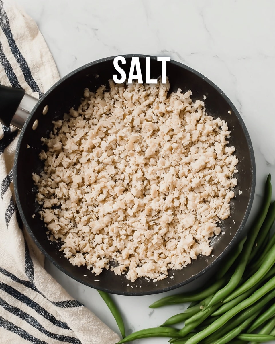 A black frying pan with small, light beige pieces of cooked food filling it completely, showing a slightly crumbly texture. The pan is placed on a white marbled surface. To the right side of the pan, there are fresh green beans laid down. A beige cloth with black stripes is peeking from the left side of the frame. The word