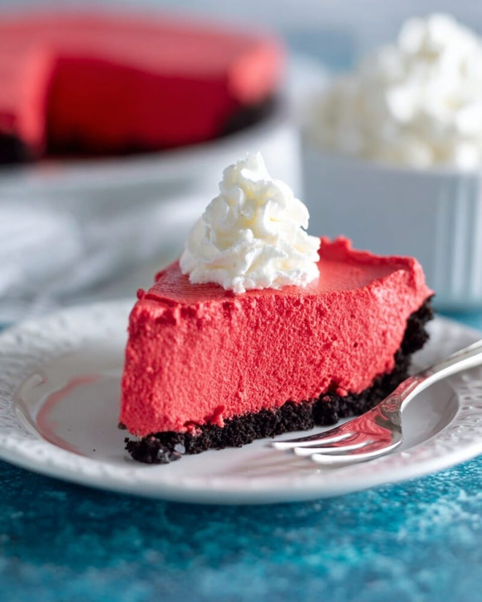 A slice of red mousse pie is shown on a white plate with a textured black crumb crust at the bottom as the first layer. Above the crust is a thick, smooth, bright red mousse layer with a light and airy texture. The slice is topped with a small dollop of white whipped cream, placed in the center. The plate is on a blue surface with a white marbled texture in the background. A fork is lying beside the slice on the plate. In the background, a white bowl filled with whipped cream and the rest of the red mousse pie in a white plate is slightly out of focus. Photo taken with an iphone --ar 4:5 --v 7