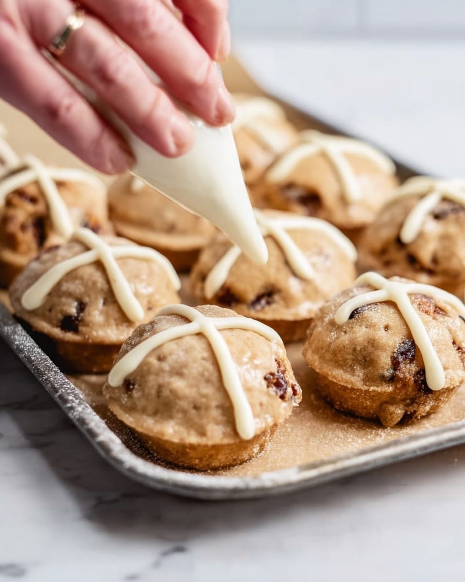 The image shows small round pastries on a metal baking tray placed on a white marbled surface. Each pastry is light brown and slightly rough in texture, with some showing darker spots where nuts or fruit pieces peek through. A woman's hand is squeezing white icing from a piping bag to create crosses on top of the pastries. The scene highlights the contrast between the warm pastry colors and the white icing being applied. Photo taken with an iphone --ar 4:5 --v 7
