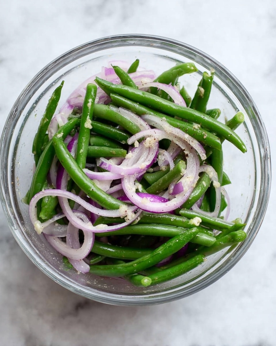 The image shows a clear glass bowl filled with fresh green beans and thinly sliced light purple onions mixed together. The green beans are vibrant and whole, layered evenly with the curved, translucent onion slices. Small bits of seasoning are visible on the vegetables, giving a subtle texture contrast. The bowl is placed on a white marbled surface. Photo taken with an iphone --ar 4:5 --v 7