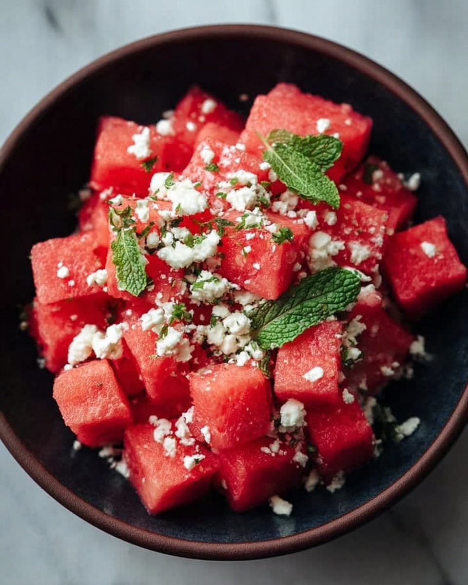 A dark bowl filled with bright red watermelon cubes forms the main layer, topped with small white crumbles of feta cheese scattered evenly across the fruit. Fresh green mint leaves are sprinkled on top, adding a pop of color and texture. The bowl sits on a white marbled surface, creating a clean and fresh background. photo taken with an iphone --ar 4:5 --v 7