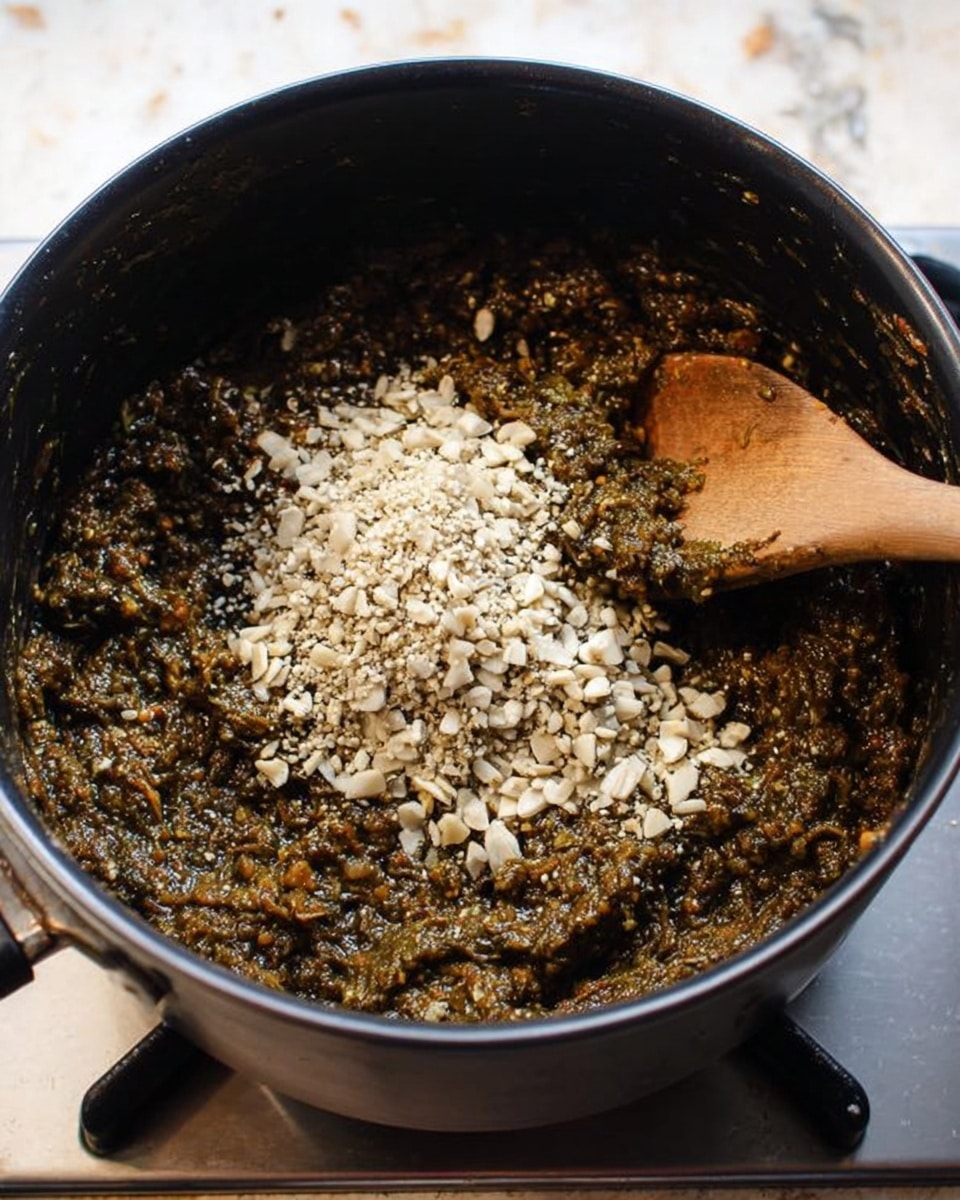 A black pot filled with a thick mixture of dark brown and greenish mashed ingredients, with a heap of small pale beige chopped pieces piled on top in the center. A wooden spoon with a medium brown handle is placed inside the pot, resting on the right side with some of the mixture sticking to its tip. The pot is positioned on a stove with a background surface changed to a white marbled texture. photo taken with an iphone --ar 4:5 --v 7