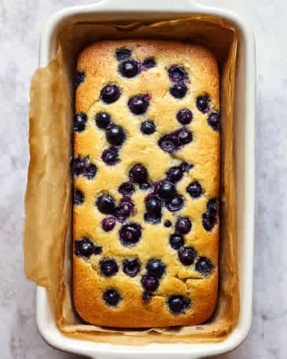 A rectangular white baking dish holds a fresh baked golden yellow cake with many blueberries scattered throughout the top surface. The cake looks soft and moist, filling the dish fully with a slightly risen center. Brown parchment paper is wrapped along the sides, peeking up above the edges of the cake. The dish sits on a white marbled surface. photo taken with an iphone --ar 4:5 --v 7