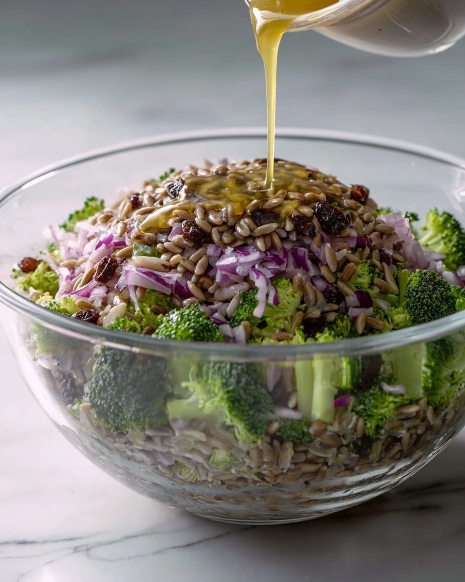 A clear glass bowl holds a fresh salad layered from the bottom with small green broccoli florets and light purple chopped onions, then topped with a thick layer of light brown sunflower seeds mixed with dark brown raisins. A golden liquid dressing is being poured over the salad, creating a shiny texture on top. The bowl sits on a white marbled surface with soft lighting highlighting the bright colors and textures of the ingredients, photo taken with an iphone --ar 4:5 --v 7