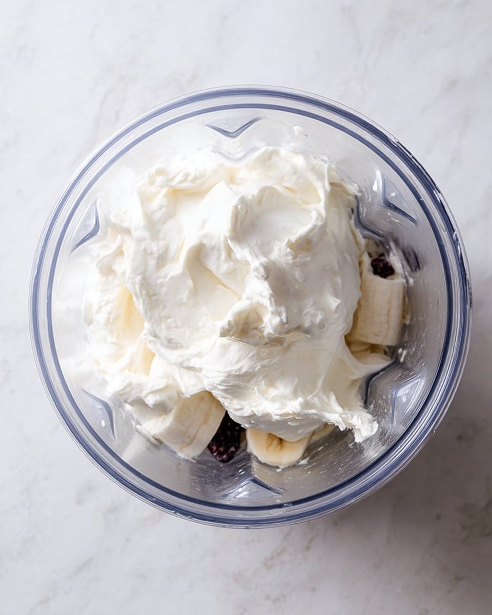 The image shows a top view of a clear blender jar filled with several layers. At the bottom, there are small pieces of white and purple fruits, possibly berries and bananas, partially visible through the jar. On top of this fruit layer, there is a thick and creamy white yogurt layer spread unevenly with soft peaks and folds. The jar is placed on a white marbled surface. Photo taken with an iphone --ar 4:5 --v 7