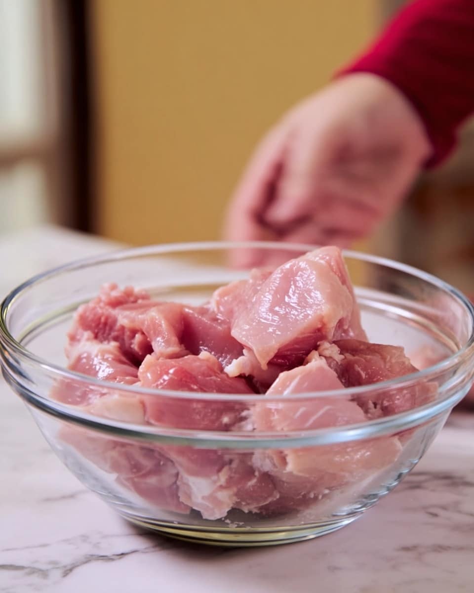 A clear glass bowl holds several raw pink chicken pieces with some white fat visible on them, stacked loosely inside the bowl. The bowl rests on a white marbled surface with some blurred background colors. A woman's hand is shown above the bowl, slightly out of focus, as if about to add or mix something photo taken with an iphone --ar 4:5 --v 7