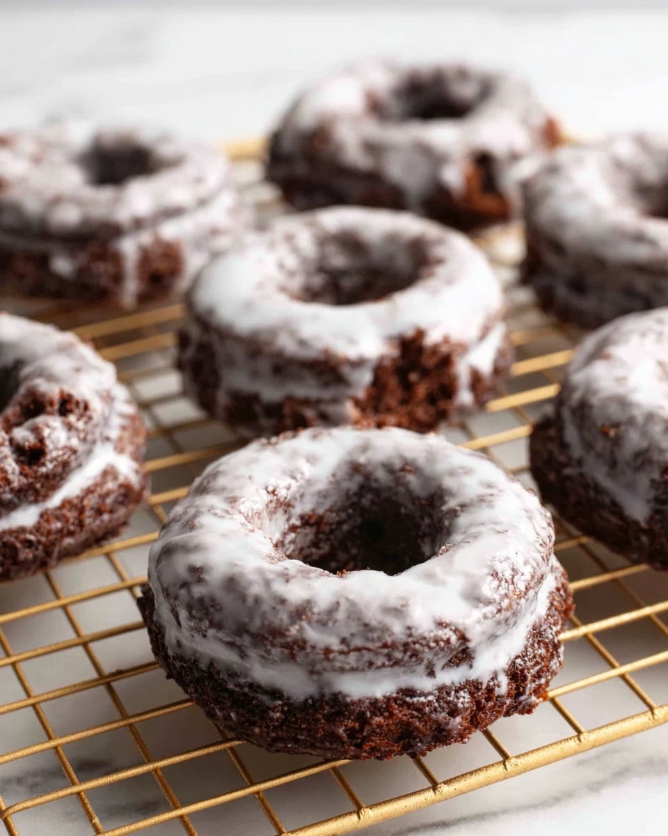 The image shows several chocolate cake donuts with a shiny white glaze coating. Each donut has a rough, crumbly texture with visible layers of dark brown cake inside, and the glaze covers the surface unevenly, pooling slightly in the donut holes. The donuts are placed on a gold cooling rack atop a white marbled surface, and the background is simple and light. Photo taken with an iphone --ar 4:5 --v 7