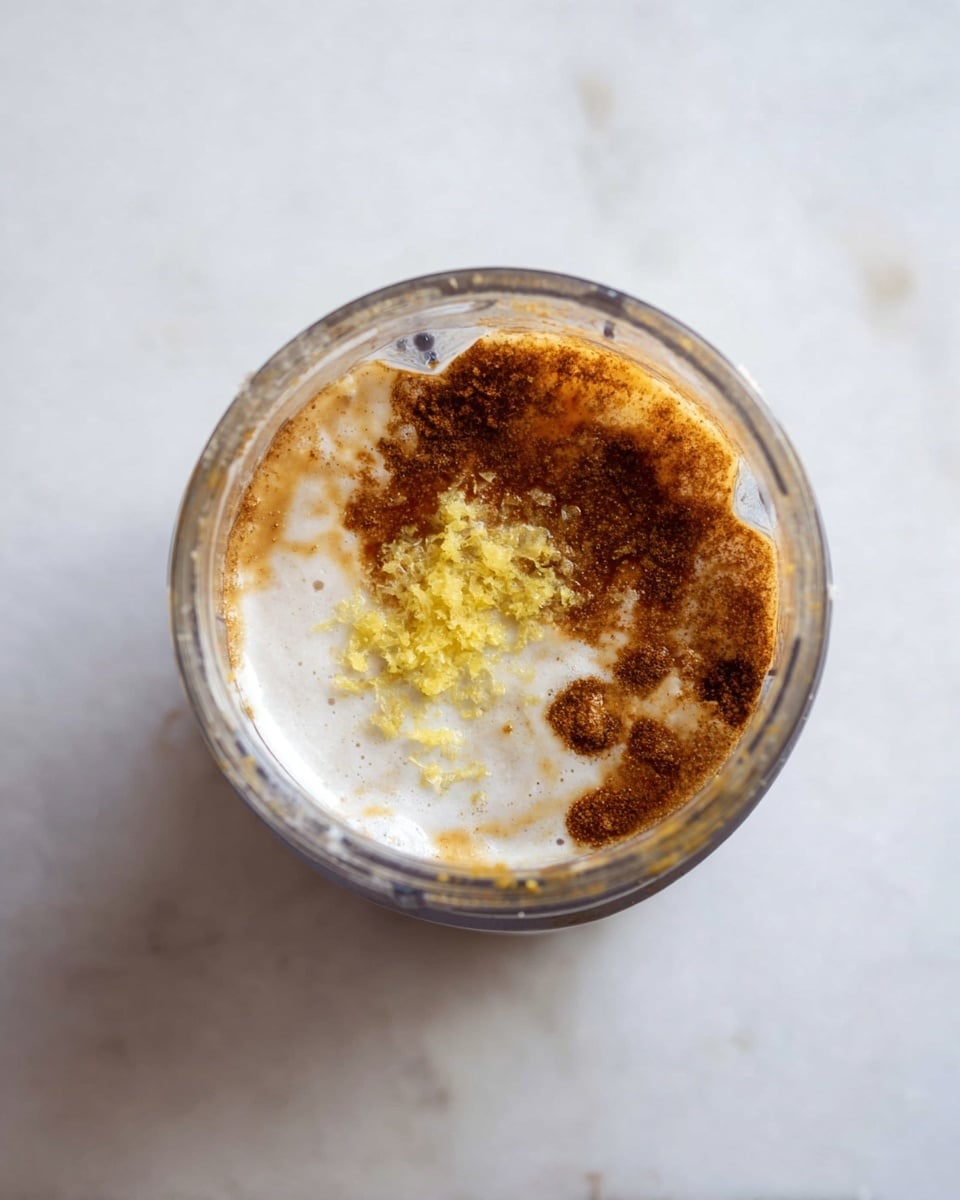 A clear blender cup is filled with several layers of ingredients from a top view. The bottom layer is a creamy, white liquid with a smooth texture. On one side, there is a light yellow zest piled in a small mound. Surrounding the zest and spreading around the cup is a darker brown, liquid layer with a slightly grainy texture. The cup sits on a white marbled surface. photo taken with an iphone --ar 4:5 --v 7