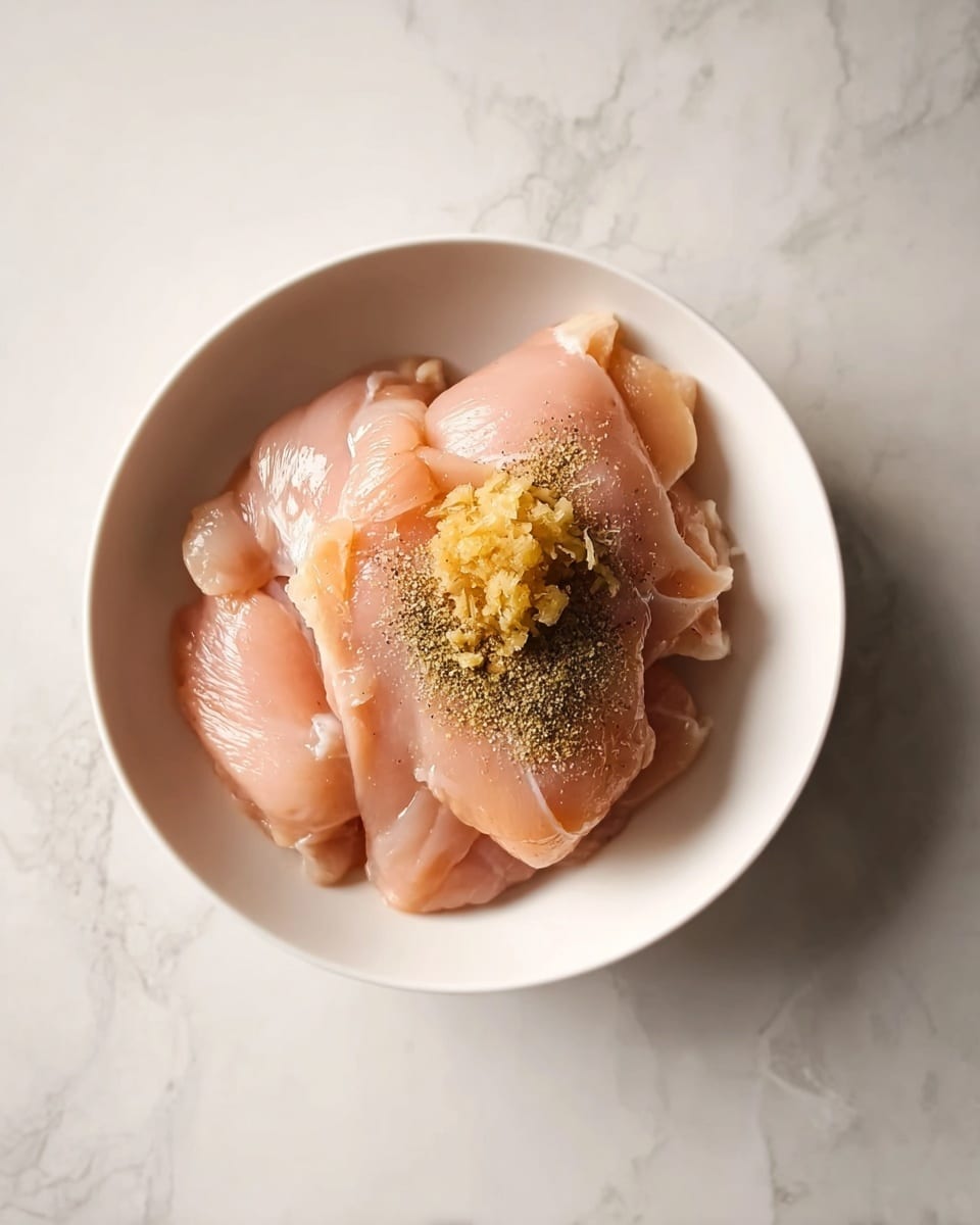 In the image, there is a white bowl on a white marbled surface, holding several layers of light pink raw chicken pieces folded over each other. On top of the chicken, there is a small pile of finely minced yellowish ginger and a sprinkle of black pepper powder. The chicken looks fresh and smooth, and the ginger adds a bit of texture, while the black pepper creates a contrast with its dark color. Photo taken with an iphone --ar 4:5 --v 7