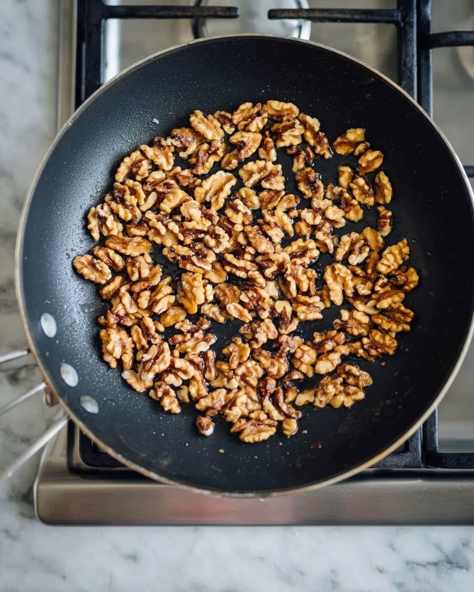 A black frying pan filled with a single layer of golden-brown walnut halves and pieces, some showing darker toasted spots, is placed on a stove with silver burners visible around the pan. The pan sits on a white marbled surface that is partially visible at the bottom of the image. photo taken with an iphone --ar 4:5 --v 7