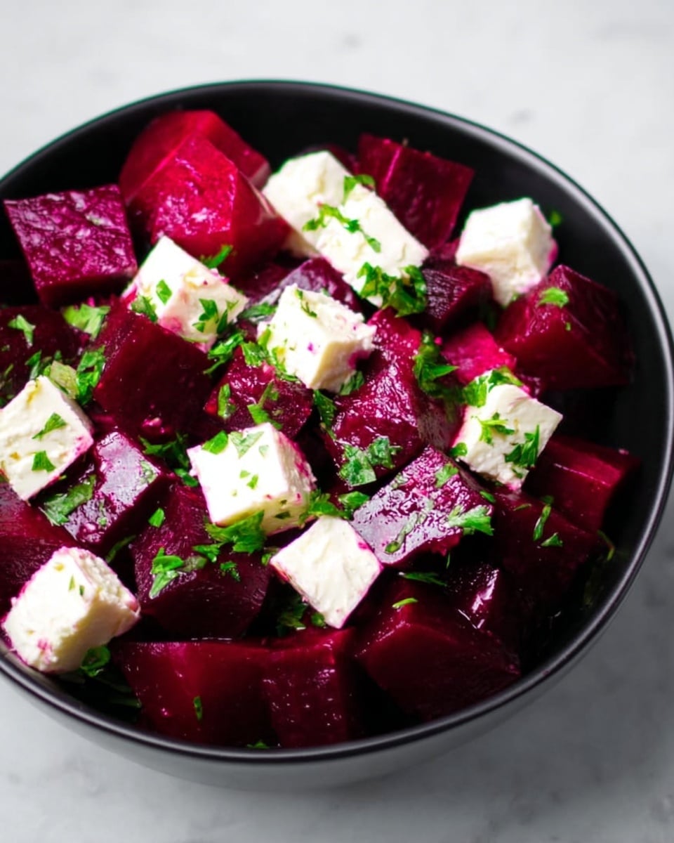 The dish shows many shiny, dark pink cubes of beetroot mixed with small white cubes of cheese on a white bowl. There are small bits of fresh green herbs sprinkled on top. The bowl sits on a white marbled surface, creating a clean and fresh look. The colors stand out with the dark pink beetroot, bright white cheese cubes, and green herbs photo taken with an iphone --ar 4:5 --v 7
