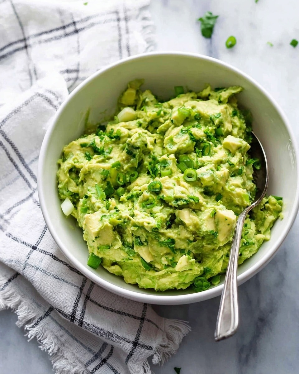 A white bowl filled with chunky avocado mash mixed with small pieces of green herbs and light green onion slices, showing a creamy yet textured mix. A silver spoon rests inside the bowl, and the bowl sits on a white marbled surface next to a white and grey checked cloth with soft loops on the edge. The colors are mainly light green and white with silver contrasts, showing a fresh and smooth combination photo taken with an iphone --ar 4:5 --v 7