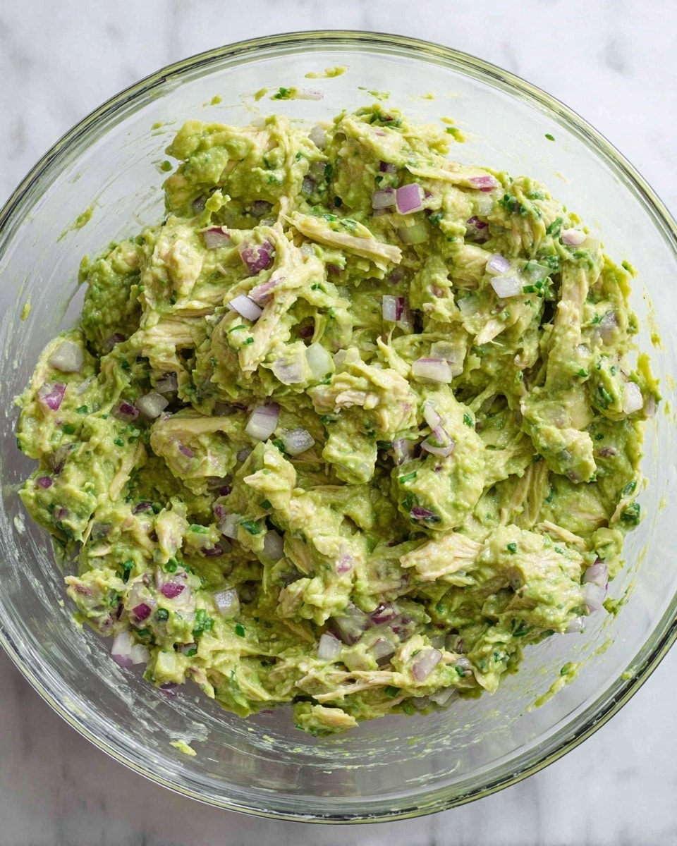 A clear glass bowl filled with a chunky green mixture that looks creamy and thick, made from mashed avocado mixed with shredded pale chicken pieces, small cubes of purple-red onion, and bits of green herbs, all mixed together giving a textured and lumpy look. The bowl is set on a white marbled surface. photo taken with an iphone --ar 4:5 --v 7