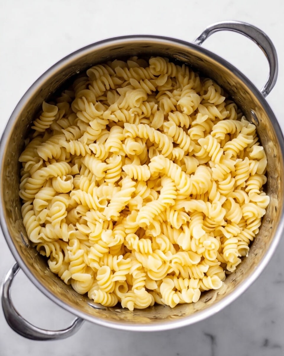 The image shows a close-up view of a pot filled with cooked spiral pasta. The pasta is light yellow in color with smooth and slightly shiny texture, filling the pot evenly. The pot is stainless steel with two metal handles on each side, set on a white marbled surface. There are no other items or layers in the image, just the plain pasta inside the pot. Photo taken with an iphone --ar 4:5 --v 7