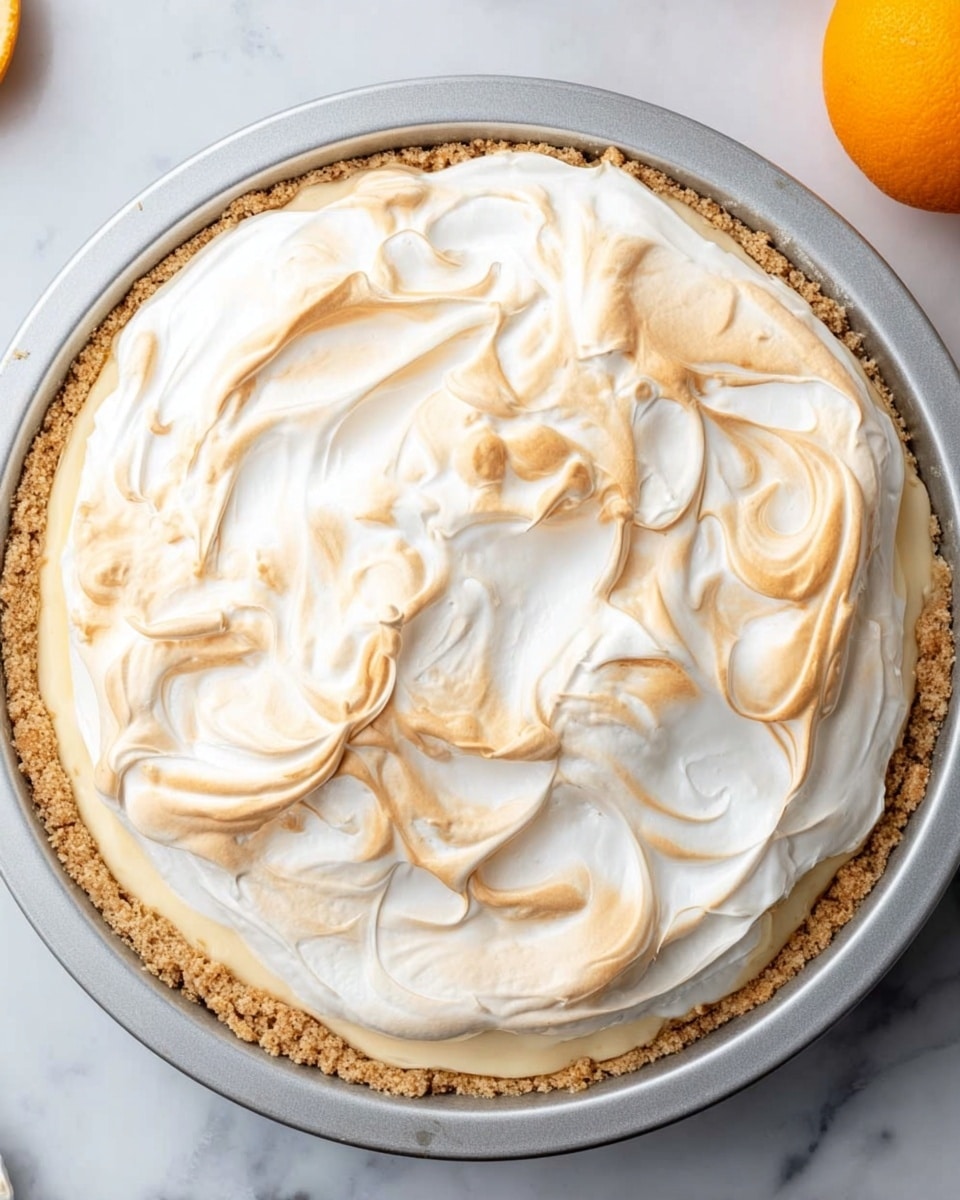 A round pie shown from above in a silver baking tin with a crumbly light brown crust at the bottom layer. The filling is creamy white and topped with a lightly toasted soft layer of meringue, showing swirls of pale golden brown and white. The meringue layer has a smooth, slightly swooped texture across the surface. The background is a white marbled texture with part of an orange fruit visible on the right edge. Photo taken with an iphone --ar 4:5 --v 7
