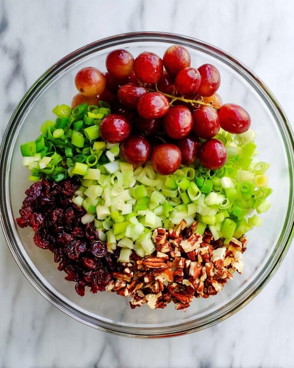 A clear glass bowl placed on a white marbled surface, filled with five separate sections of ingredients arranged in a circle. One section has halved red grapes showing smooth deep red and light green interiors, next to a pile of chopped fresh green onions with bright green and white colors. Adjacent to these is a heap of finely diced pale green celery. Next to the celery is a cluster of small dark red dried cranberries, and beside them is a pile of chopped brown nuts with crunchy texture and some whitish pieces mixed in. photo taken with an iphone --ar 4:5 --v 7
