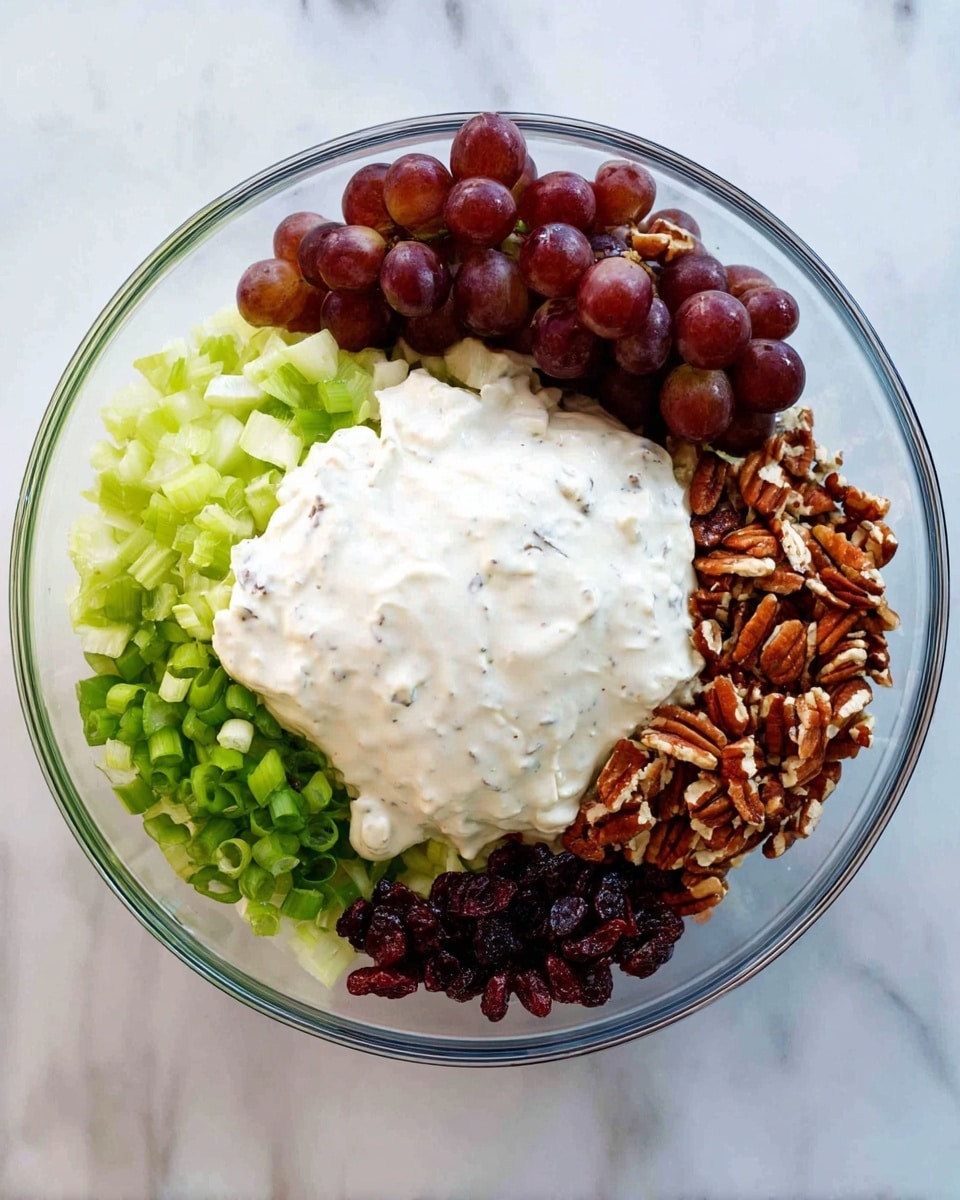 A clear glass bowl sits on a white marbled surface, filled with layers of fresh ingredients arranged in separate sections. There is a cluster of whole purple grapes on one side, next to a pile of finely chopped light green celery. A large dollop of creamy white sauce with a smooth texture and tiny black specks is placed in the center, partially covering the other layers. Around the sauce, there are chopped green onions with bright green color and chopped pecans with a mix of dark brown and tan rough textures. At the bottom edge, there are dark red dried cranberries. Each ingredient is distinct and neatly placed, showing a colorful and fresh mix. photo taken with an iphone --ar 4:5 --v 7