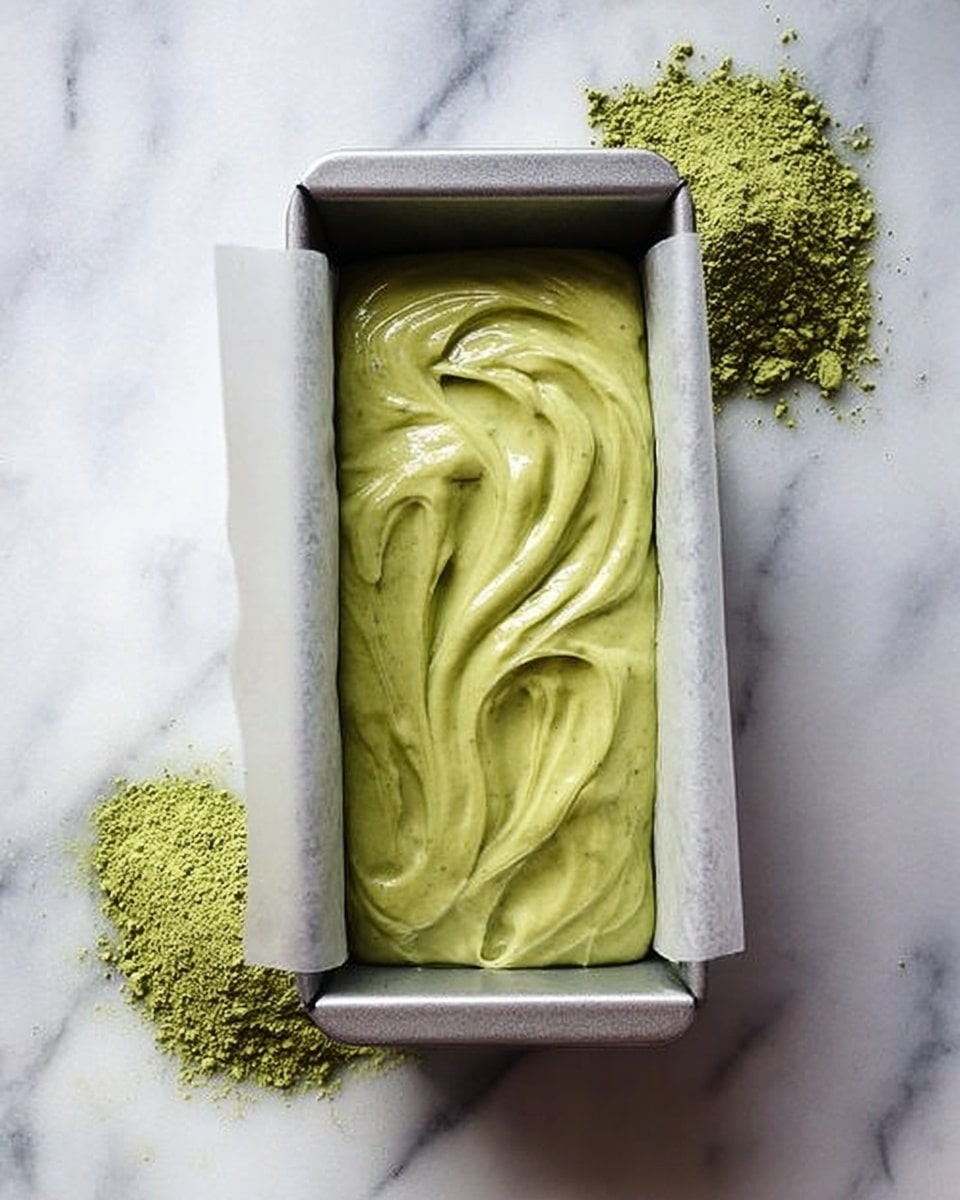 The image shows a loaf pan lined with parchment paper, filled with smooth green batter that has soft swirls on its surface. The pan sits on a white marbled surface, and there are two small piles of green powder nearby, one on each side of the pan. The batter looks thick and creamy with a slightly shiny texture. photo taken with an iphone --ar 4:5 --v 7