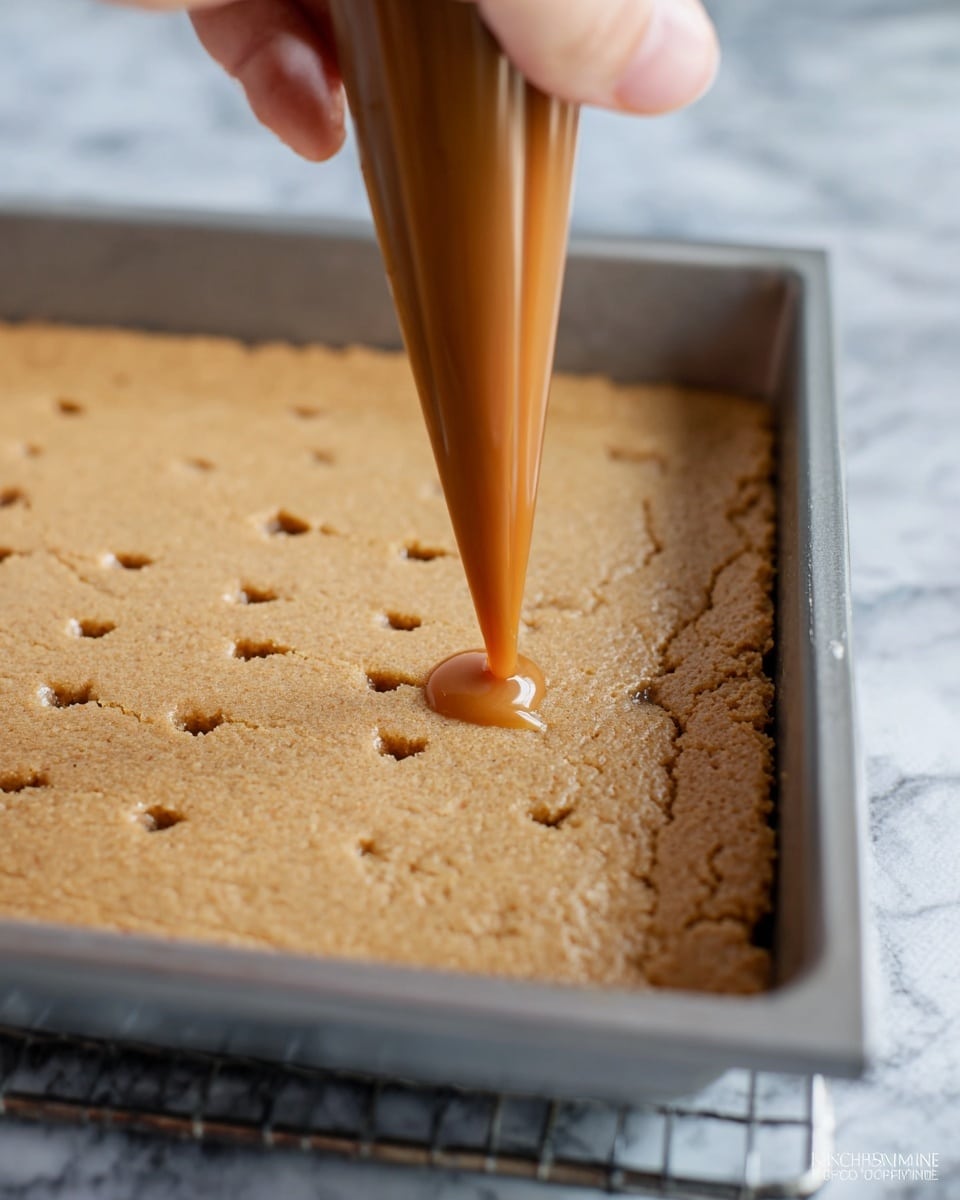 A close-up image shows a woman's hand holding a piping bag filled with smooth caramel sauce, gently squeezing it to fill small holes evenly spaced in a large single layer of light brown baked cake or bar base inside a metal baking tray. The textured surface of the baked layer has scattered tiny cracks and crevices, while the background shows a soft focus of the white marbled kitchen surface. photo taken with an iphone --ar 4:5 --v 7