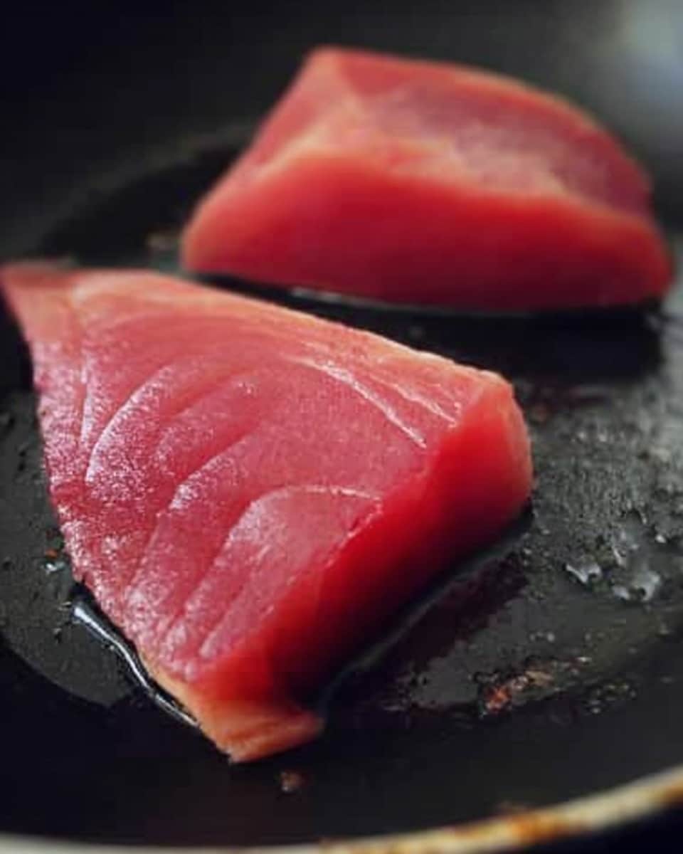 Two raw tuna pieces in a dark frying pan, with a deep red color and smooth texture. The tuna pieces are thick and neatly shaped, showing different shades of red and pink. The pan surface is a little shiny, reflecting soft light. The edges of the tuna pieces are clean and slightly curved. The background is a white marbled texture. photo taken with an iphone --ar 4:5 --v 7
