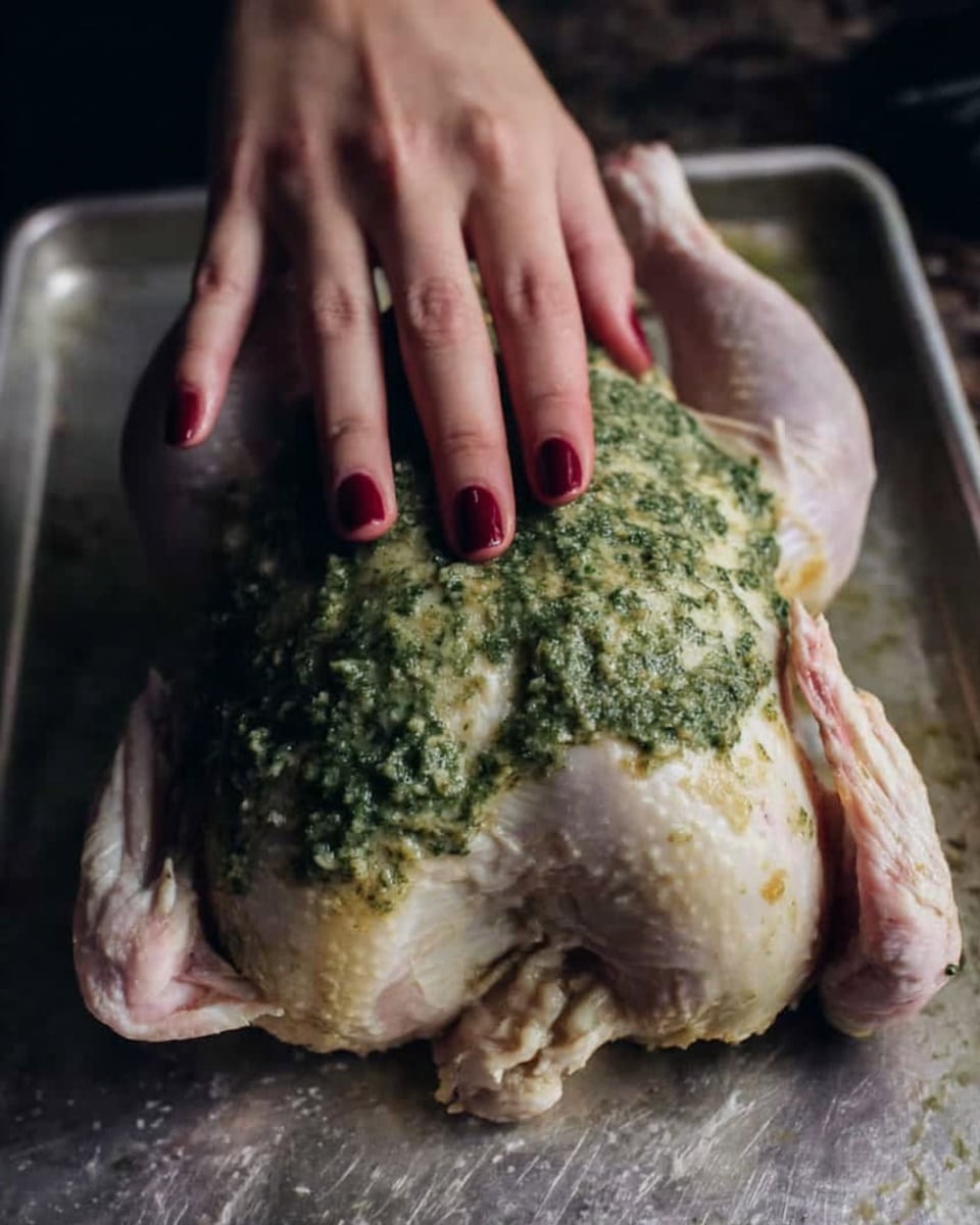 A raw whole chicken lies on a metal tray, skin pale and slightly shiny. On top of the chicken’s back is a layer of green herb paste with a rough texture, spread unevenly. A woman's hand with dark red painted nails is pressing down gently on the chicken near the herb layer, her fingers slightly splayed. The chicken’s legs and wings are relaxed to the sides. The scene is lit softly, highlighting the contrast between the pale chicken skin and the rich green of the herb paste, with the cold metal tray underneath. photo taken with an iphone --ar 4:5 --v 7