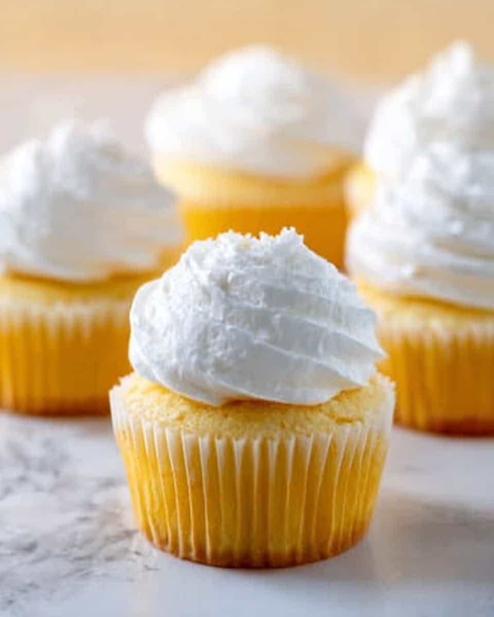 The image shows five yellow cupcakes with a soft texture, each topped with a thick, smooth layer of white frosting that forms a rounded mound on top. The cupcakes are placed on a white marbled surface with bright lighting that brings out the creamy and fluffy look of the frosting and the moist texture of the cake layers beneath it. The background is softly blurred, making the cupcakes the clear focus. photo taken with an iphone --ar 4:5 --v 7