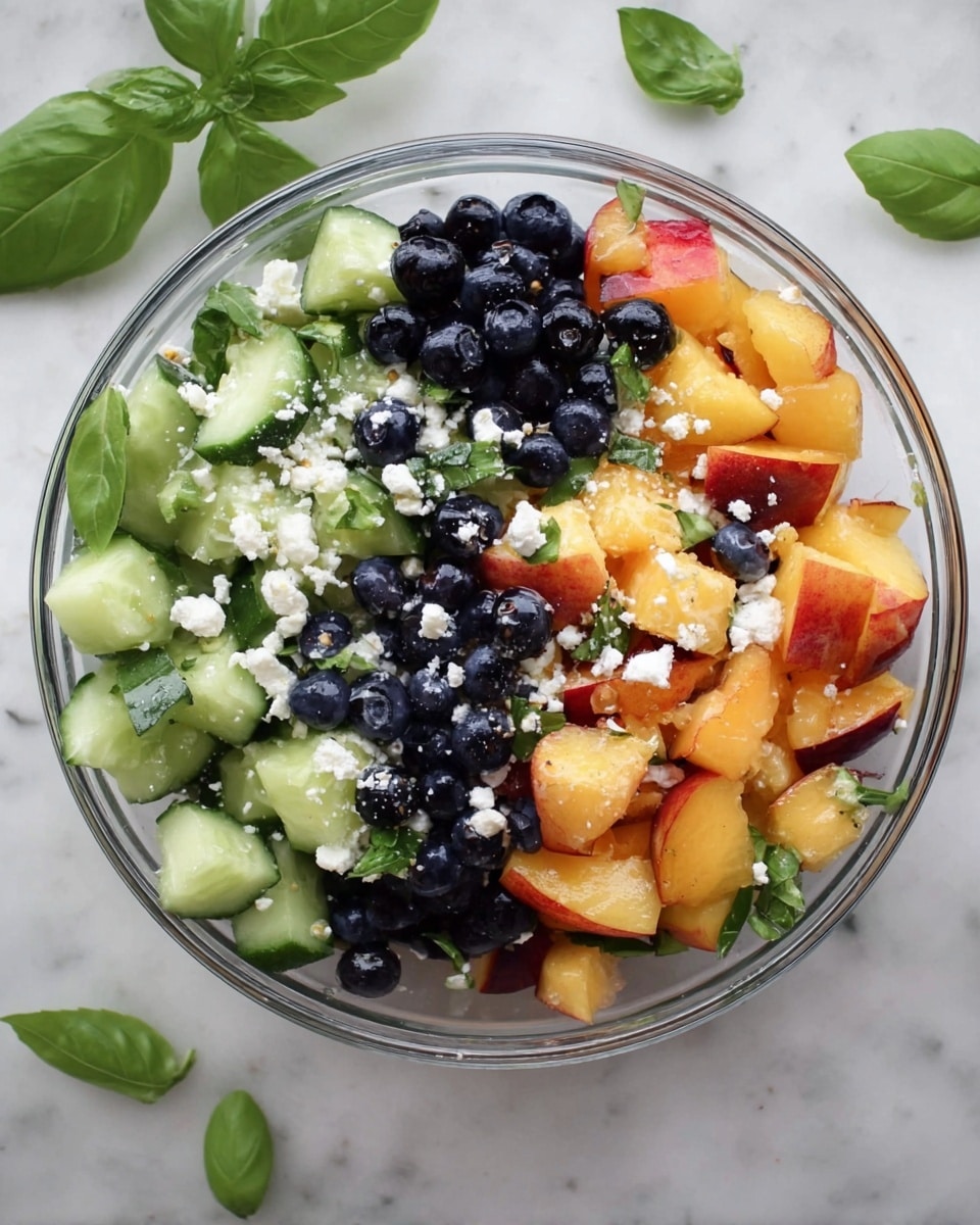 The image shows a clear bowl filled with a colorful salad that has four main layers visible. The bottom layer is fresh, green cucumber slices with smooth edges. The second layer has bright orange chunks of peach or nectarine with red skin spots. The third layer is made up of small, round, dark blue blueberries scattered evenly. The top layer is sprinkled with white crumbles of cheese, adding a soft texture. Around the salad, fresh green basil leaves are placed for decoration. The bowl is placed on a white marbled surface. photo taken with an iphone --ar 4:5 --v 7