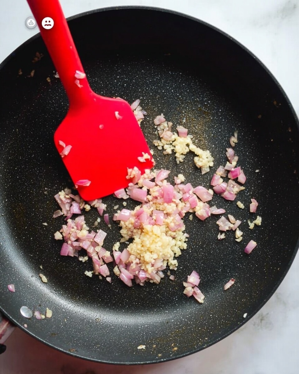 A close-up view of a black non-stick pan on a white marbled surface, with small pieces of pink shallots and light yellow minced garlic being stirred by a red silicone spatula. The shallots are scattered in a circular pattern around the center of the pan, where the garlic is slightly clumped together, showing a soft and lightly cooked texture. The scene captures the early stage of cooking, with the ingredients gently sizzling and releasing aroma. Photo taken with an iphone --ar 4:5 --v 7
