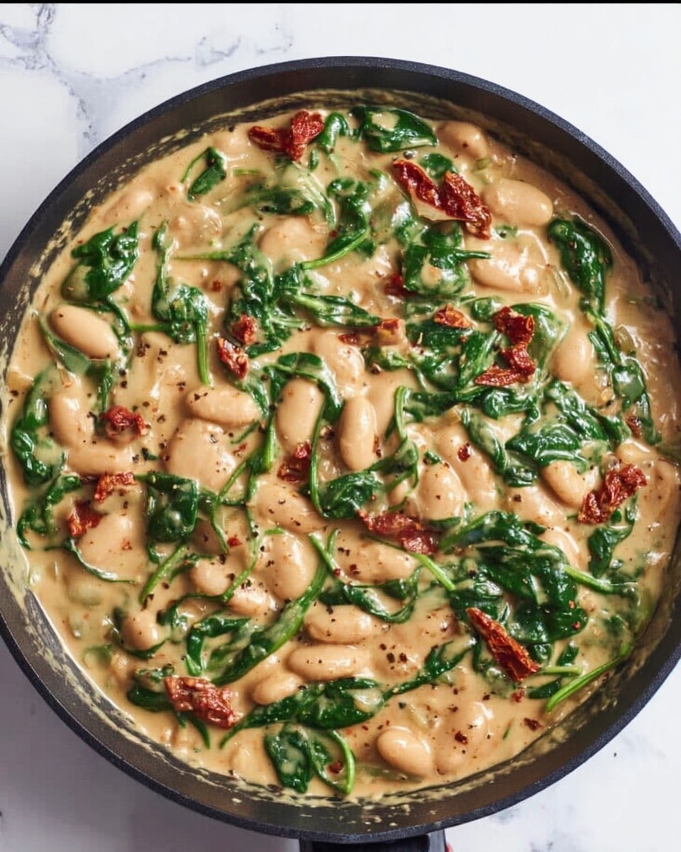 The image shows a black skillet filled with a creamy dish made of large white beans, bright green spinach leaves, and small bits of reddish sun-dried tomatoes, all mixed in a thick light beige sauce with some visible black pepper flakes. The beans and spinach are spread evenly throughout the sauce, creating a rich texture, with the sun-dried tomato pieces scattered randomly across the surface. The skillet is on a white marbled surface. photo taken with an iphone --ar 4:5 --v 7
