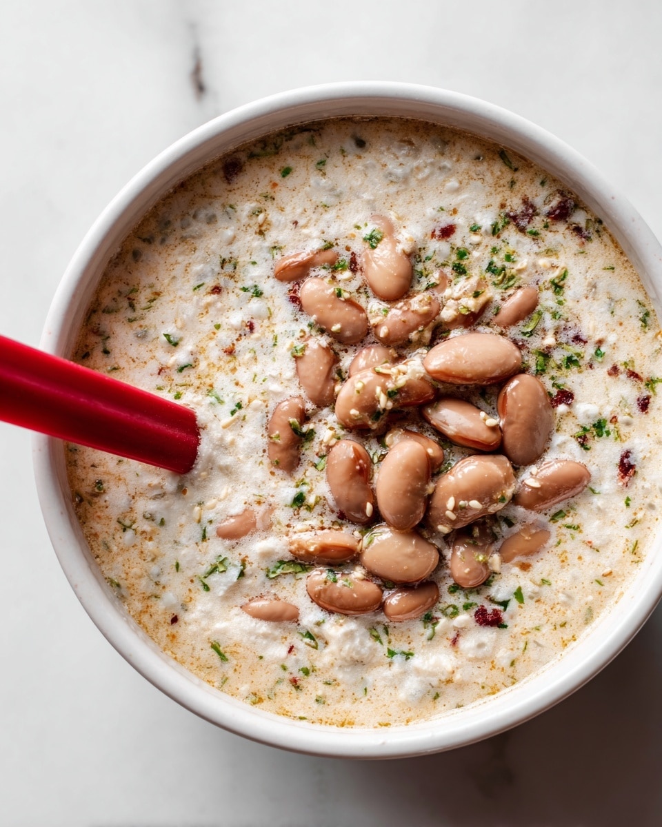 A close-up image of a white bowl filled with a creamy mixture that has small bits of herbs and seeds spread throughout. On top of the creamy base, there is a light brown layer of large beans piled in the center. A red silicone spoon is partially visible on the left side, immersed in the mixture. The bowl sits on a white marbled surface. Photo taken with an iphone --ar 4:5 --v 7