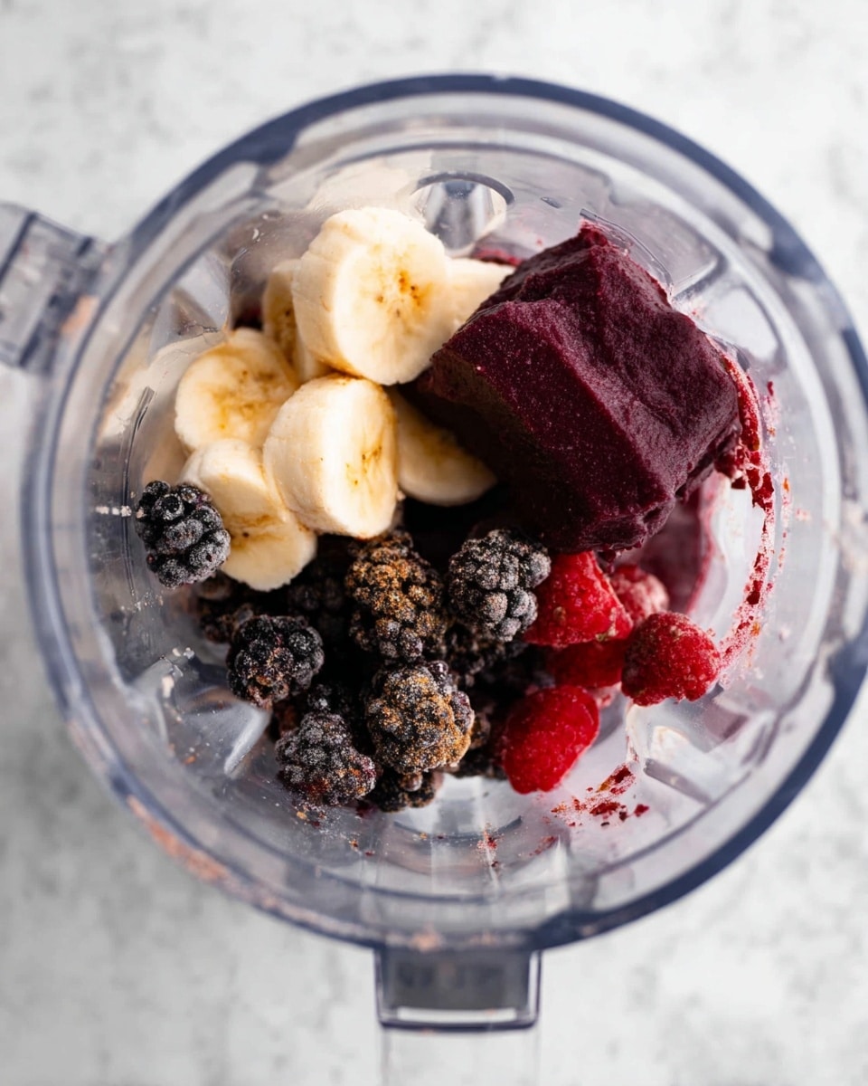 Inside a clear blender container, there is a mix of several layers of fruit. At the bottom center, soft white banana pieces slightly browned at the edges. Around the banana, there are dark purple-black frozen berries clustered together, including some whole round blackberries and small, bright red raspberries. Toward the top right, there is a larger block of dark purple fruit with a smooth but frozen texture. The blender rests on a white marbled surface. Photo taken with an iphone --ar 4:5 --v 7