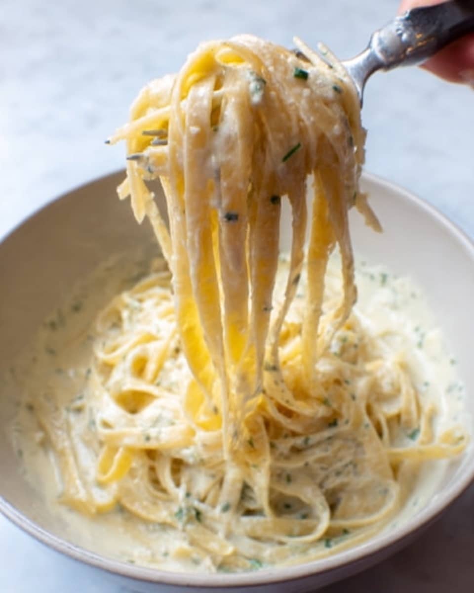 A close-up of a creamy pasta dish in a white bowl, with thick fettuccine noodles lifted by a woman's hand using a pasta fork. The creamy sauce is pale yellow with a smooth texture, speckled with small green herbs, covering the noodles evenly. The sauce looks rich and slightly thick, clinging to the pasta strands, while some noodles hang down in loose strands, showing their soft texture. The background is a white marbled surface, bright and clean, with natural light coming from behind. photo taken with an iphone --ar 4:5 --v 7