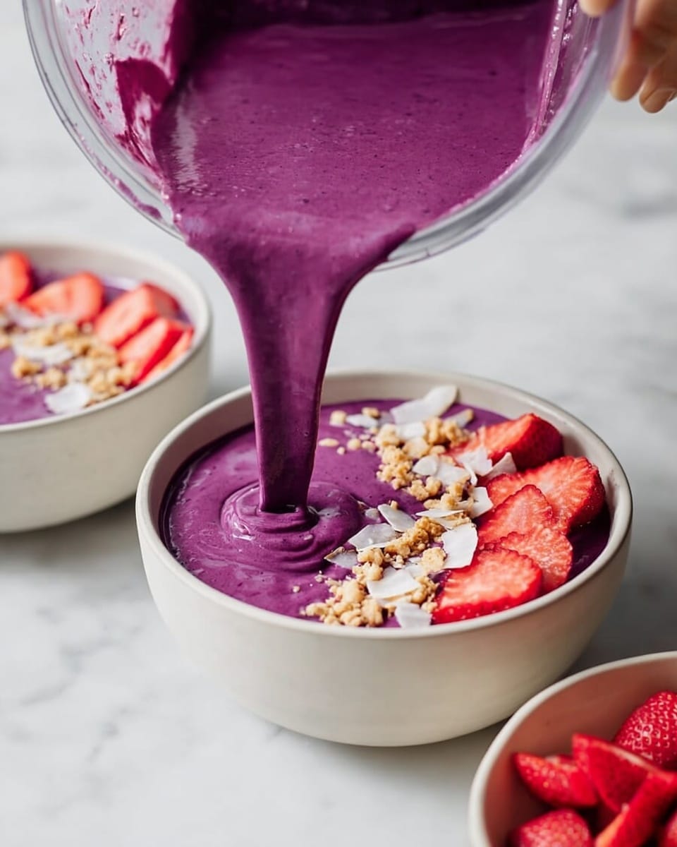 A white bowl is filled with a thick, bright purple smoothie being poured from a blender by a woman's hand visible in the top right corner. Behind it, a second white bowl holds a finished smoothie layered with toppings: the purple smoothie base is topped with sliced strawberries, light beige granola clusters, and small pieces of coconut flakes, arranged neatly across the surface. In the bottom right corner, part of a third white bowl filled with fresh, sliced strawberries is visible. All bowls sit on a white marbled surface. Photo taken with an iphone --ar 4:5 --v 7