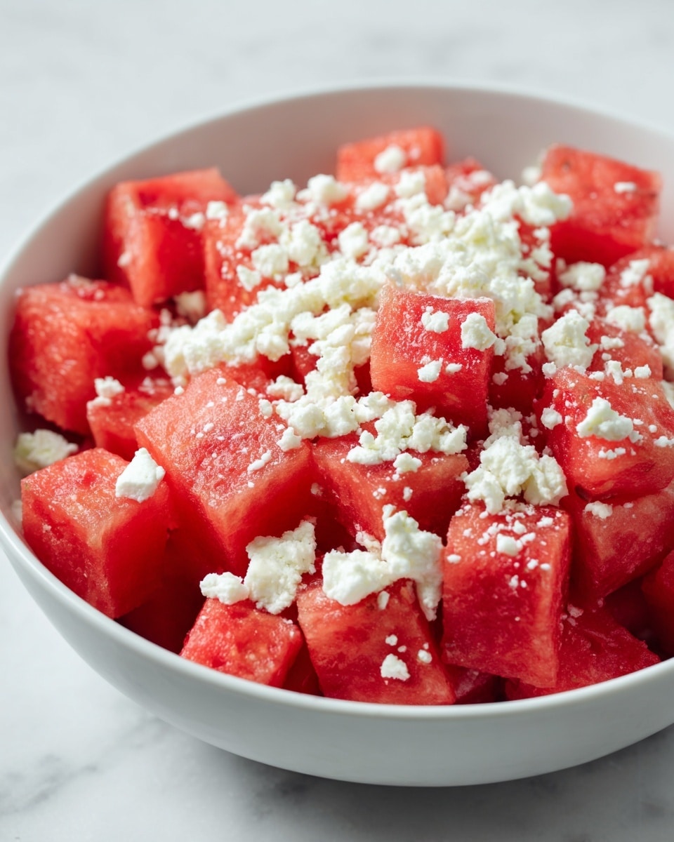 A white bowl filled with many red watermelon cubes inside. On top of the watermelon, there is a layer of white crumbly cheese scattered evenly. The surface under the bowl is white marble, giving a clean and fresh look. The colors mainly are red from the watermelon and white from the cheese and bowl, creating a bright and fresh feeling. photo taken with an iphone --ar 4:5 --v 7