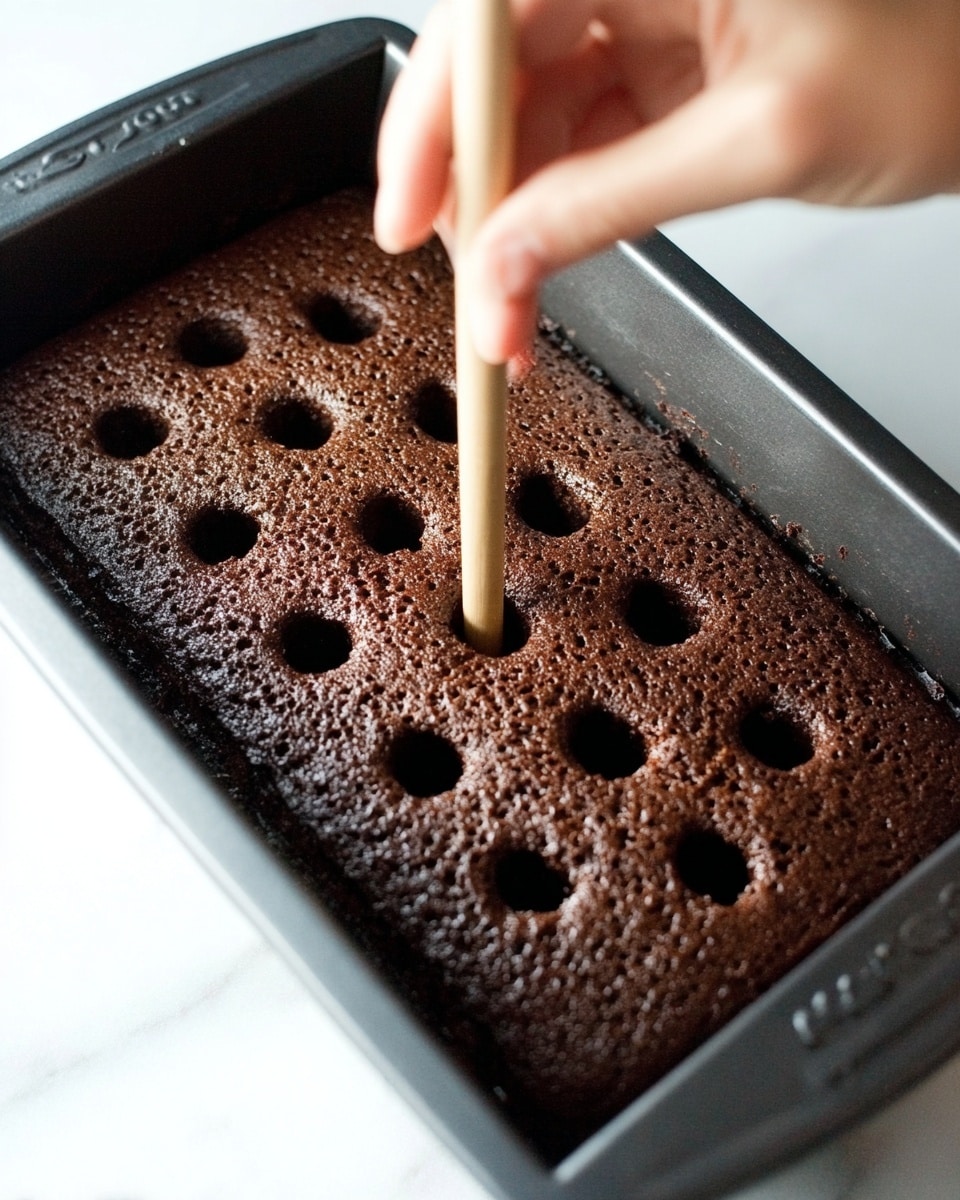 The image shows a close-up of a chocolate cake in a rectangular pan with several evenly spaced holes poked into the top layer. The cake has a rich, dark brown color with a slightly bumpy texture on the surface. A woman's hand is holding a wooden stick and pressing it into one of the holes to create another small opening. The pan is black and rests on a white marbled surface, which adds a clean and bright contrast to the dark cake. photo taken with an iphone --ar 4:5 --v 7