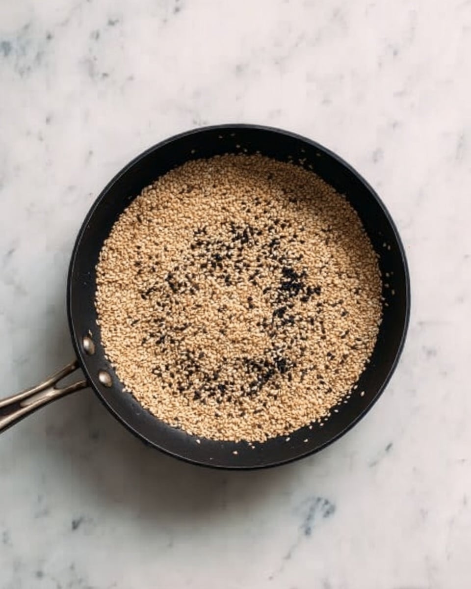 A black pan filled with a single even layer of light brown toasted sesame seeds, some darker toasted bits scattered throughout, resting on a white marbled surface photo taken with an iphone --ar 4:5 --v 7