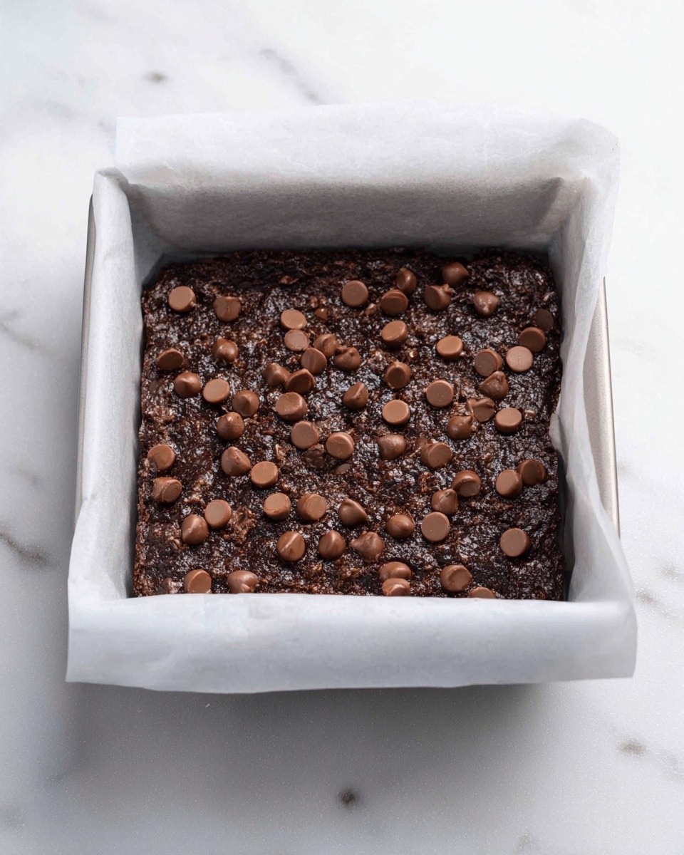 A rectangular baking pan lined with white parchment paper holds a single thick layer of dark brown chocolate dessert with a coarse texture, topped with scattered small round chocolate chips in a lighter brown shade. The edges of the parchment paper rise slightly above the dessert, and the background is a white marbled surface. photo taken with an iphone --ar 4:5 --v 7