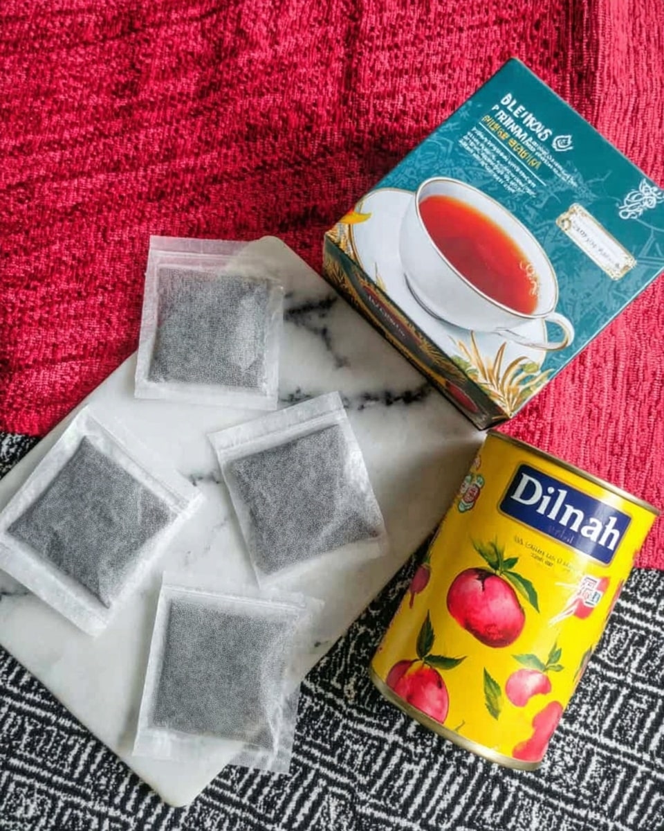 The image shows a group of six flat tea bags arranged on a white marbled surface, next to a box of Dilmah Premium tea featuring a design with a white cup of tea on the front. To the right of the tea box is a yellow tin can with red fruit graphics, placed on the same white marbled surface. The background includes a textured black and white geometric cloth and a red fabric. photo taken with an iphone --ar 4:5 --v 7