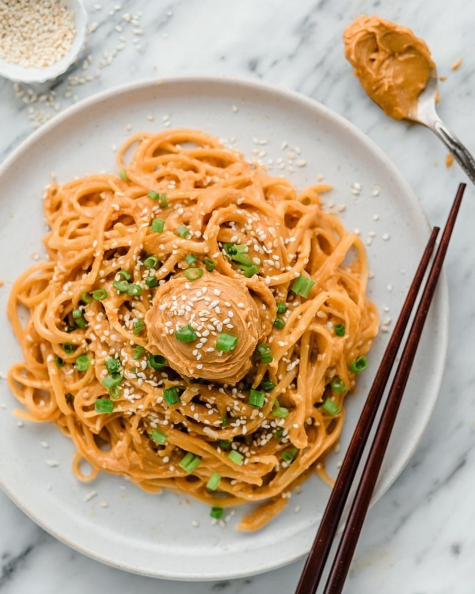 A white plate full of cooked noodles with a creamy orange sauce, topped in the center with a dollop of smooth peanut butter, sprinkled with white sesame seeds and chopped green onions. The noodles are tangled loosely around the peanut butter. Two dark brown chopsticks rest along the right edge of the plate. The plate sits on a white marbled surface, with a spoonful of peanut butter and some scattered sesame seeds nearby. Photo taken with an iphone --ar 4:5 --v 7