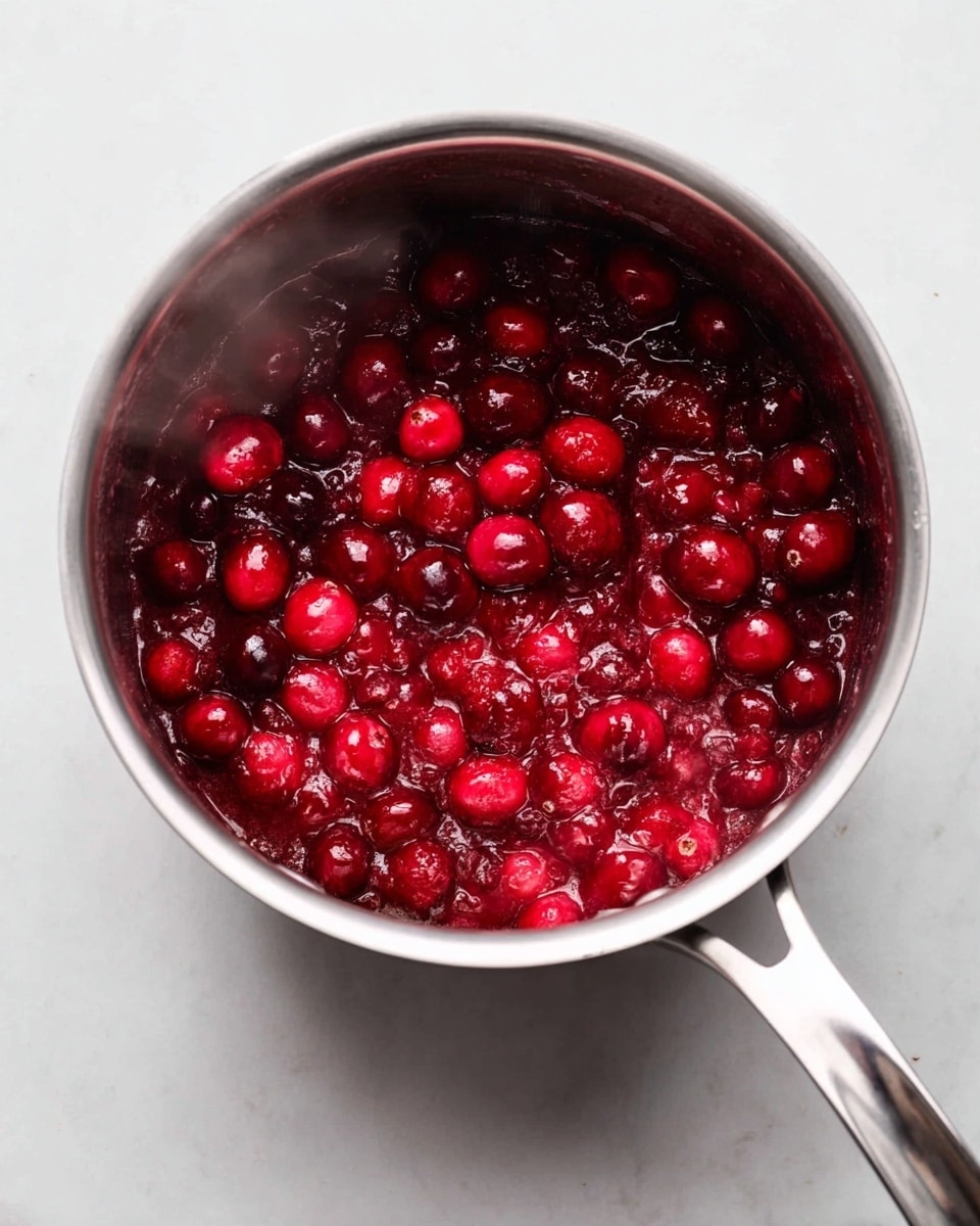 A shiny stainless steel pot is filled with bright red cranberries cooking down into a thick sauce. The cranberries are plump and shiny, some whole and some broken, creating a mix of round shapes and soft, jam-like texture. The pot sits on a white marbled surface, and steam rises slightly from the warm sauce. The handle of the pot extends out to the right. The colors are mainly deep reds with a few hints of darker shades where the cranberries have softened, giving a rich, vibrant look. Photo taken with an iphone --ar 4:5 --v 7